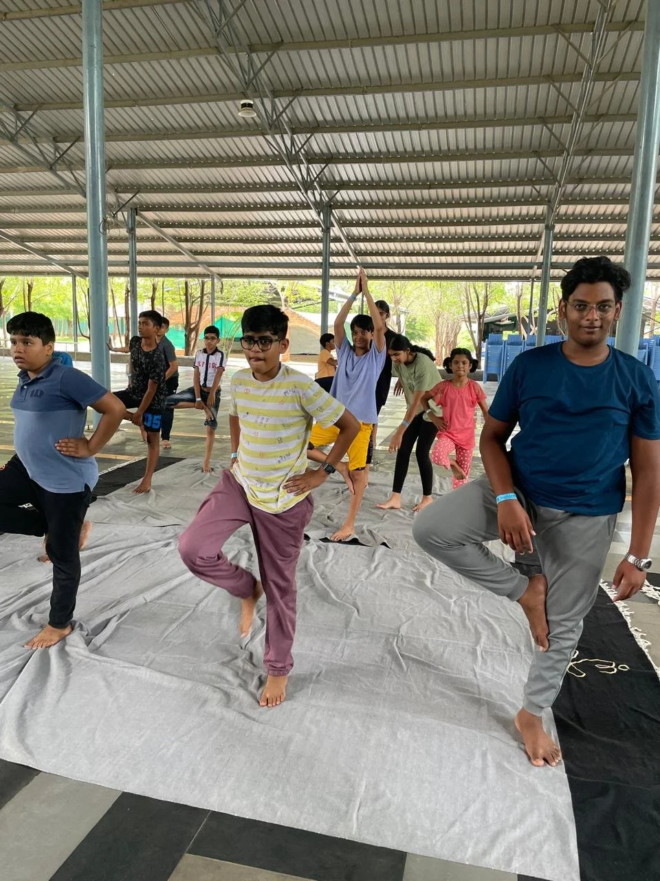 Group of children and teenagers practicing yoga or dance on a large cloth in a covered outdoor space.