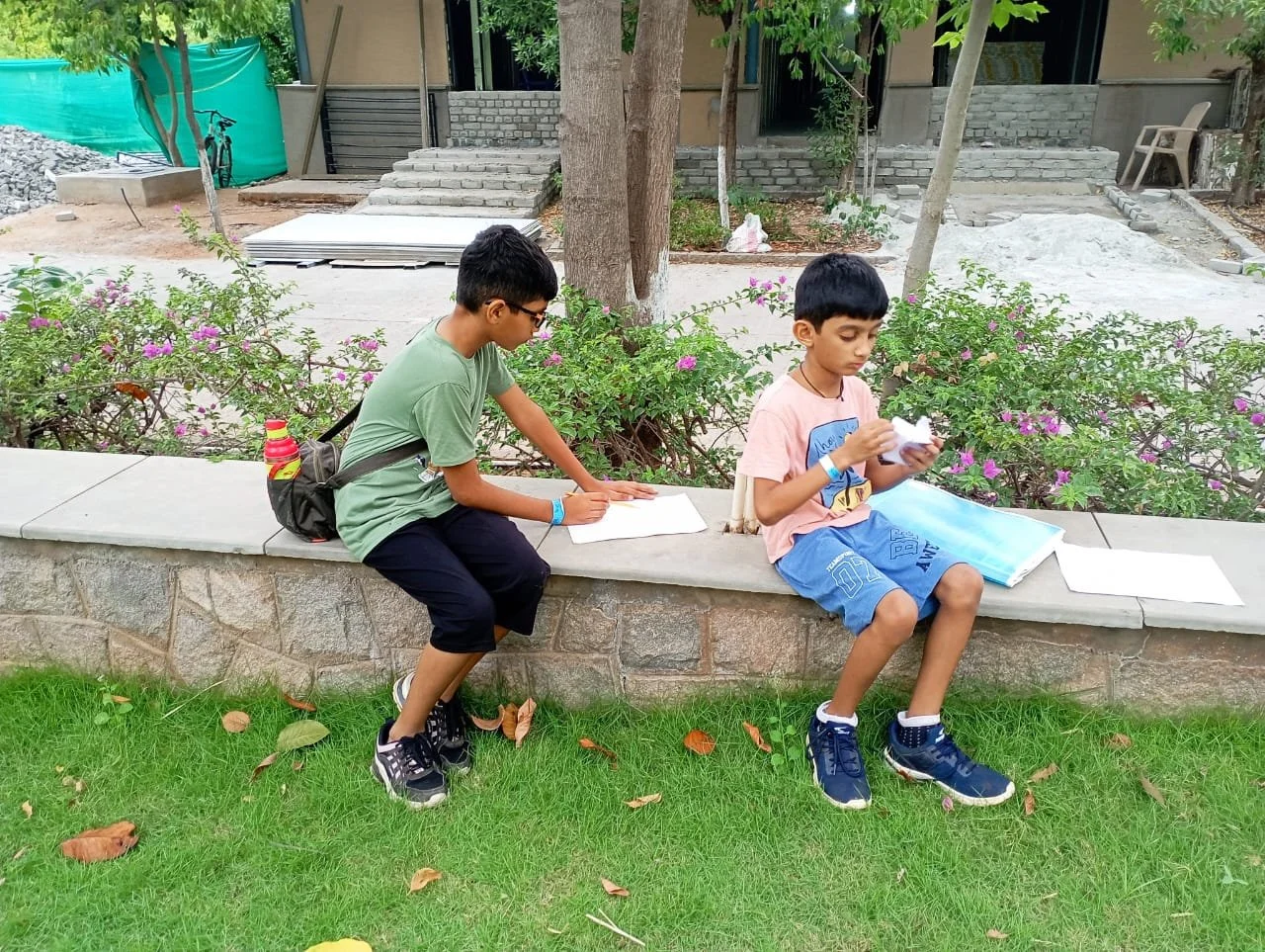 Two young boys sit on a stone ledge in a park, reading and looking at papers. One boy is wearing glasses, a green shirt, and black shorts, while the other is in a pink shirt and blue shorts. There are trees, flowering bushes, and a building in the ba