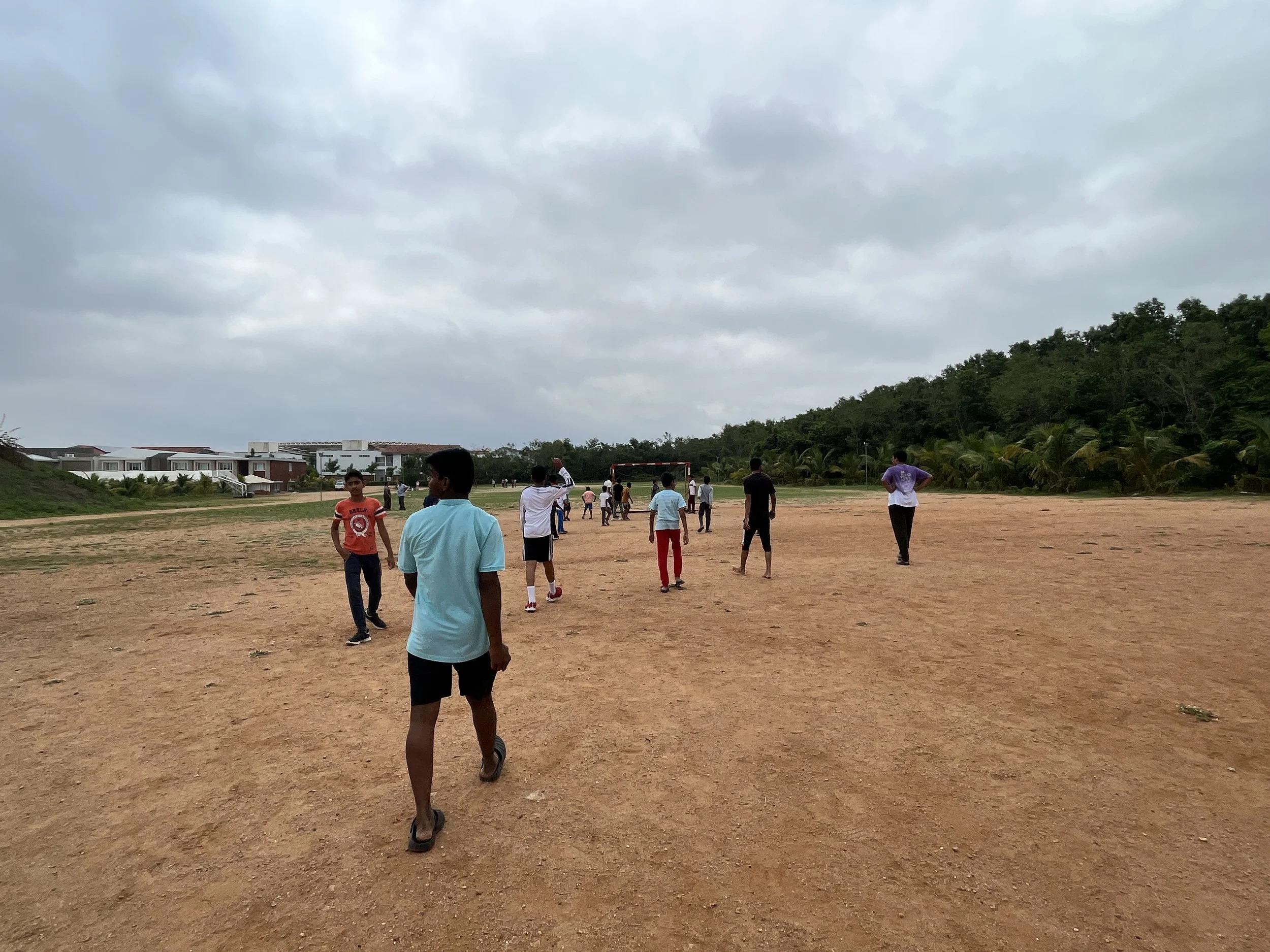 Group of children and teenagers playing or walking on a large dirt field under cloudy sky, with some houses and green trees in the background.