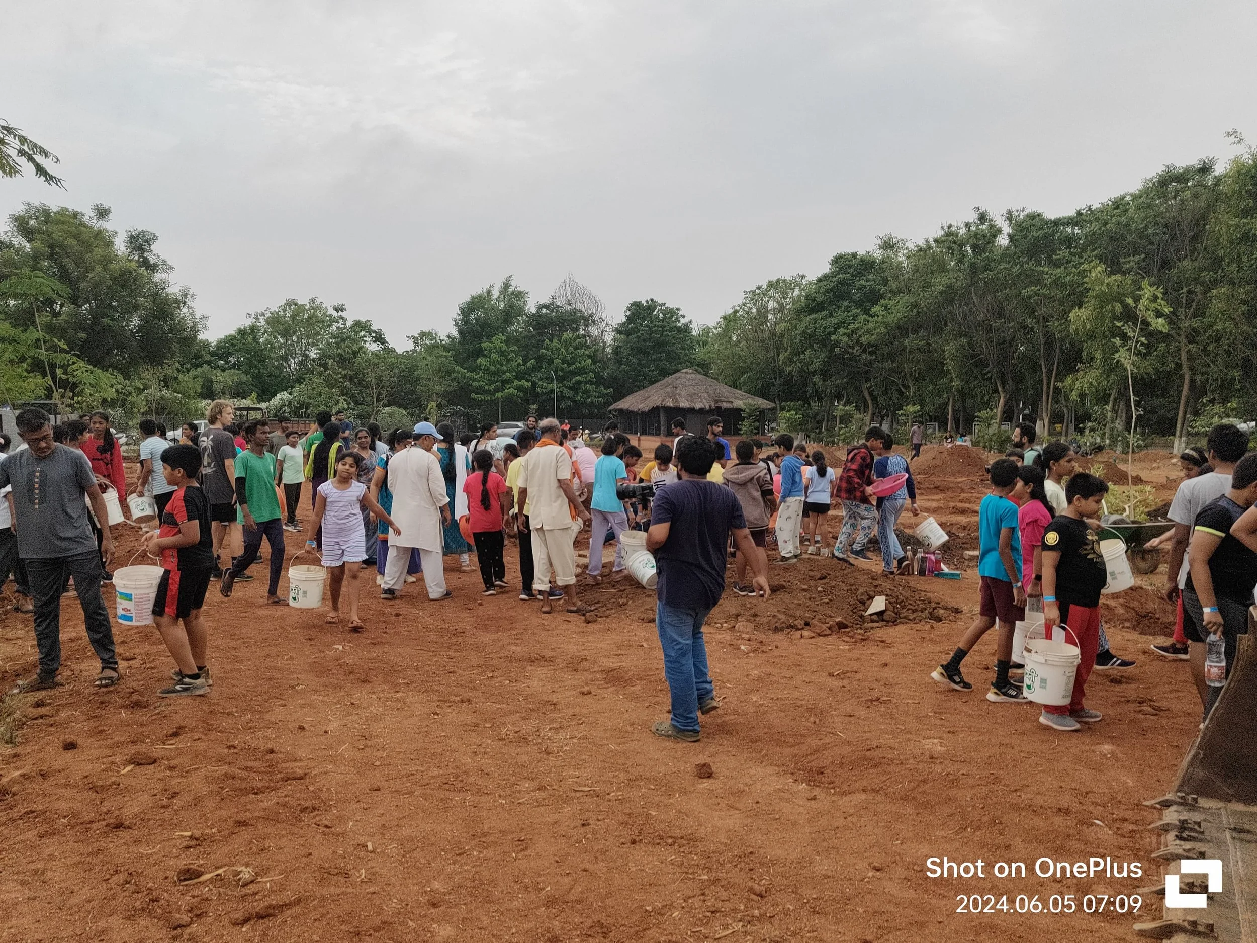 A large group of children and adults gathered outdoors on a dirt area, some carrying buckets, possibly for planting or community work, with trees and a small hut in the background.
