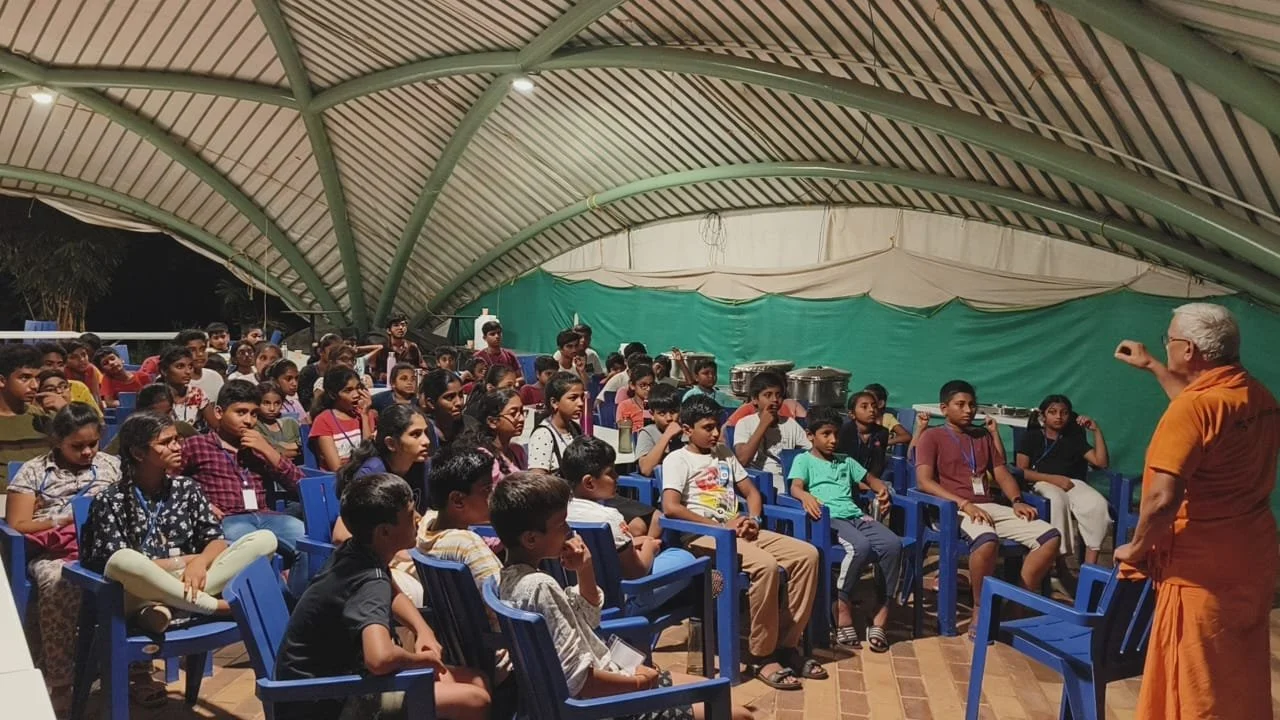A teacher or speaker addressing a large group of children seated under a large, curved, tent-like structure with green and white fabric. The children are sitting in blue chairs, listening attentively.