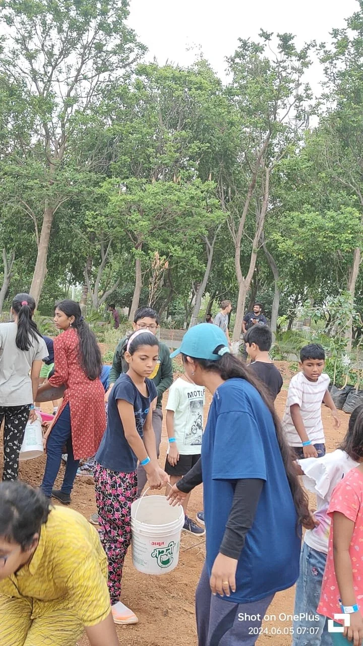 Group of children and adults planting trees or working in a garden outdoors on a cloudy day, surrounded by trees and greenery.