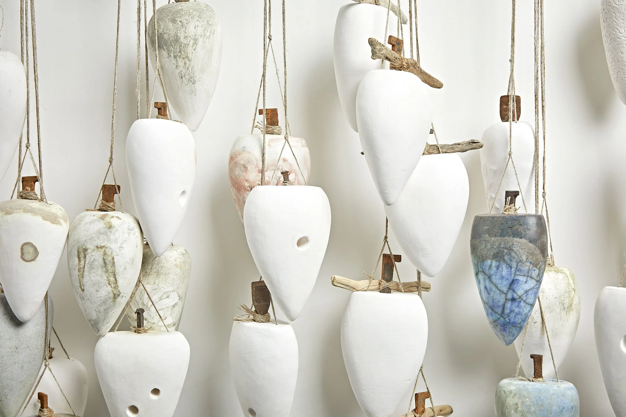 A wall of hanging ceramic vases and sculptures in various shapes and colors, suspended with ropes and wooden accents.