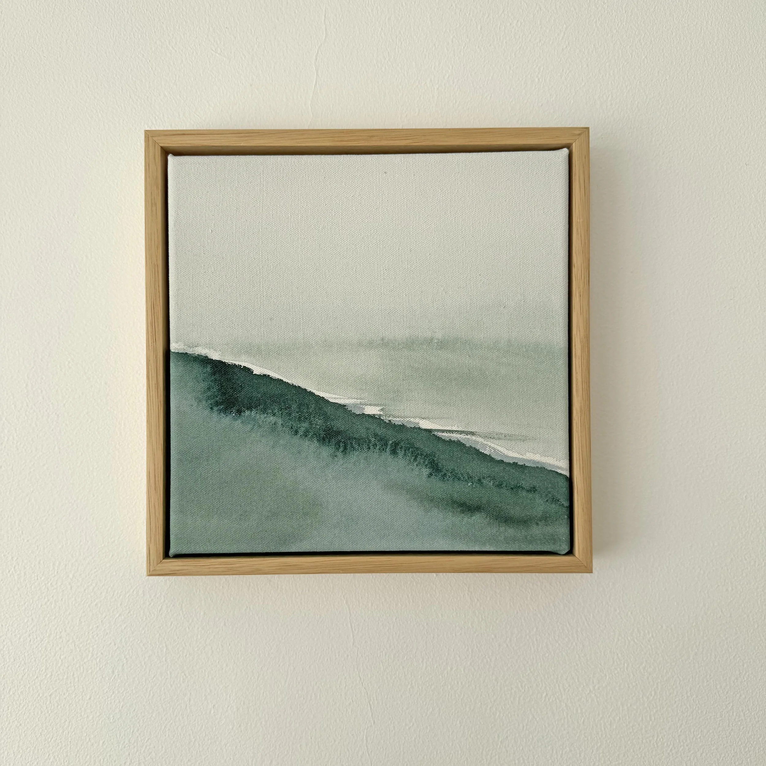 A framed piece of artwork with a landscape painting featuring hills and water, hanging on a cream-colored wall.