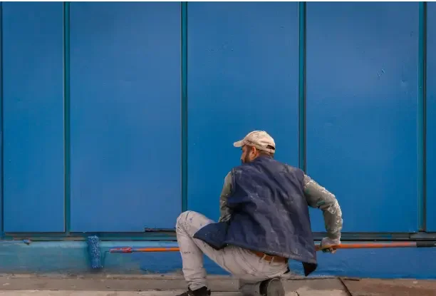 Painter applying blue exterior latex paint to an outdoor wall with a roller.