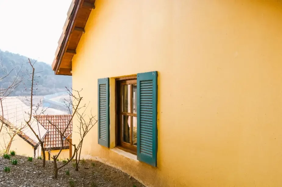 Yellow stucco home exterior with a wooden window and blue shutters overlooking a hillside neighborhood.
