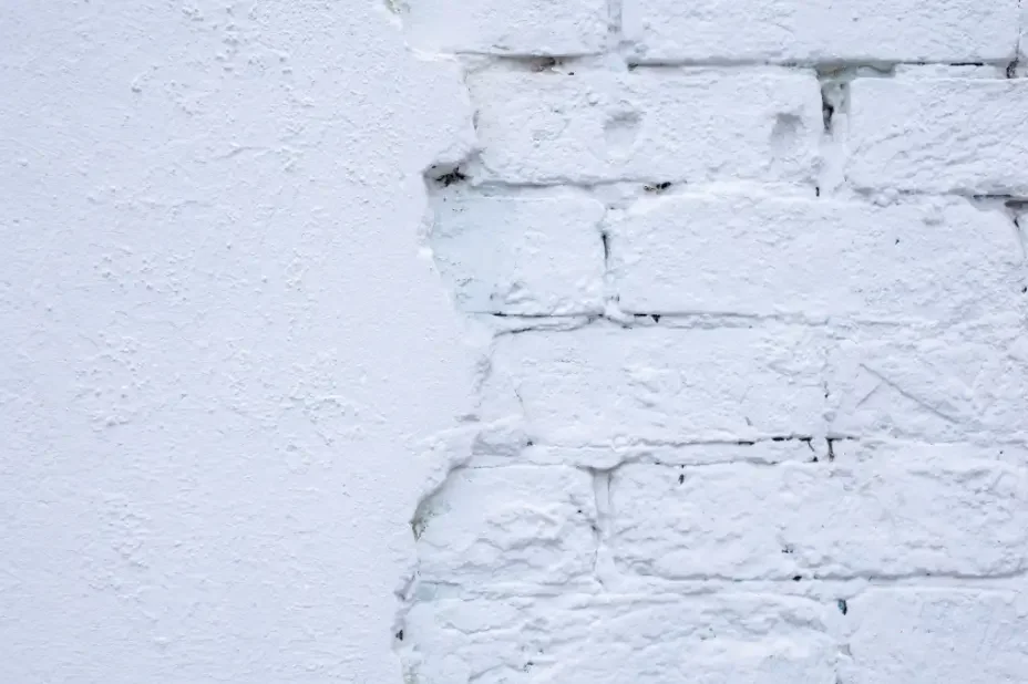 White painted brick wall with damaged stucco, exposed masonry, cracks, and uneven patches showing signs of surface failure.