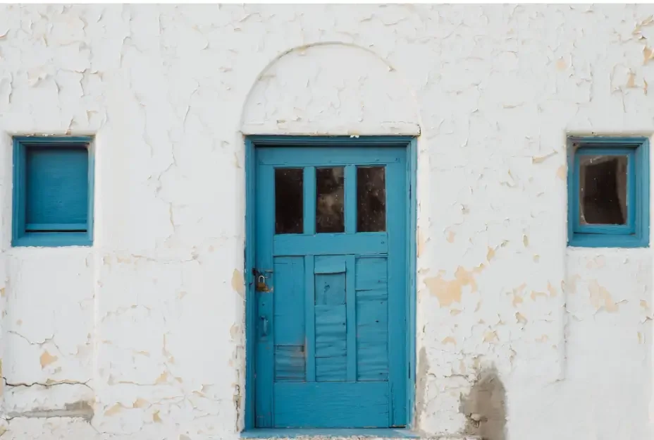 Weathered white stucco exterior with peeling paint, a blue front door, and blue window frames showing signs it is time to repaint.