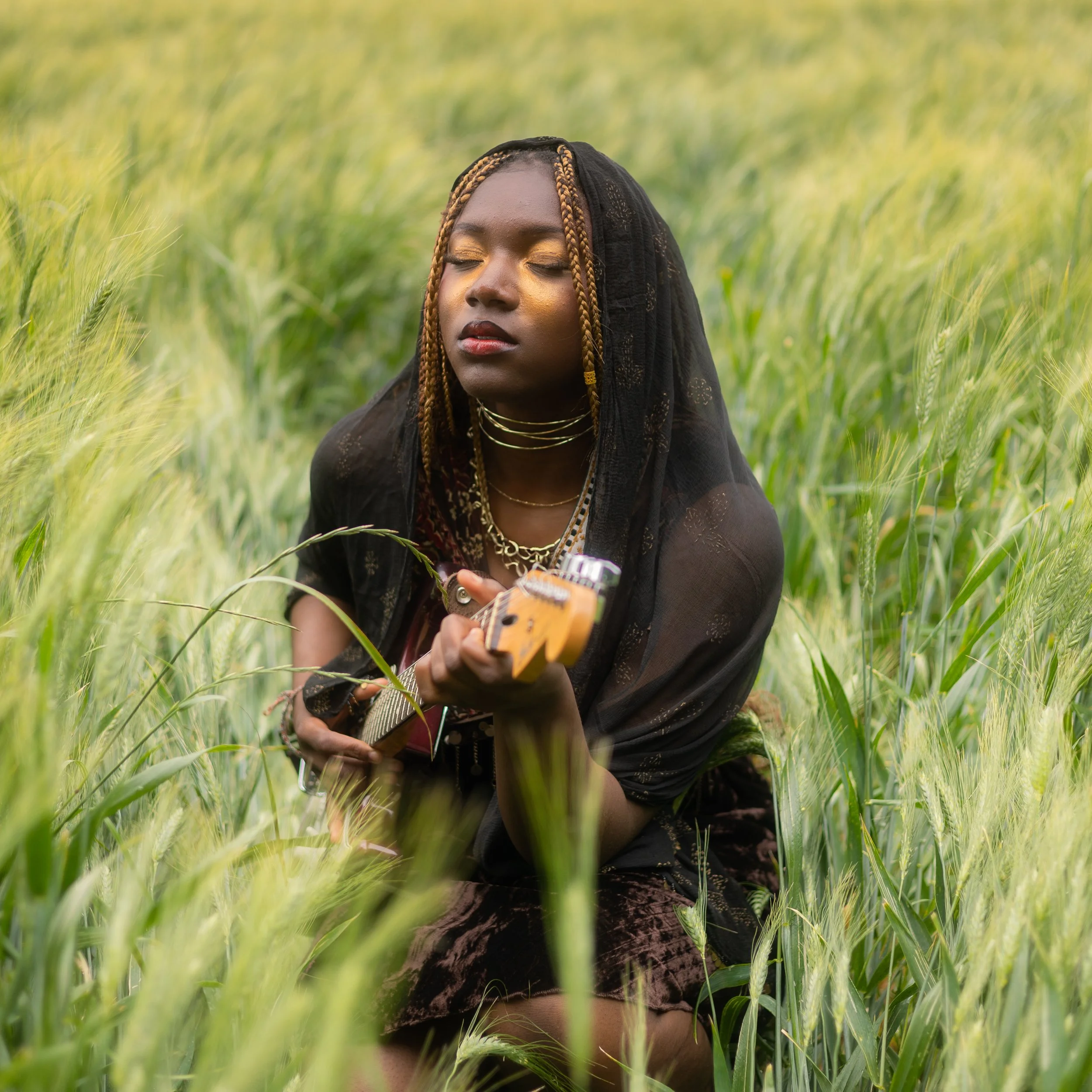 Femme noire jouant de la guitare dans un champ de blé vert avec ses yeux fermés.