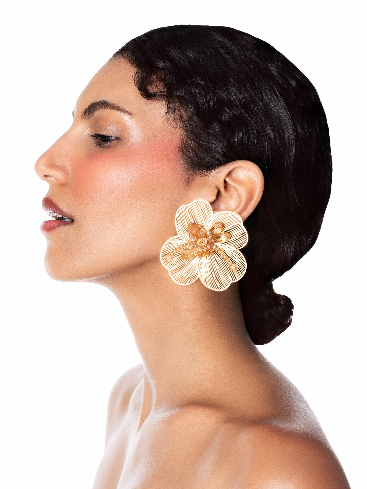 Une femme avec des cheveux noirs courts porte une grande boucle d'oreille en forme de fleur dorée avec des détails floraux.