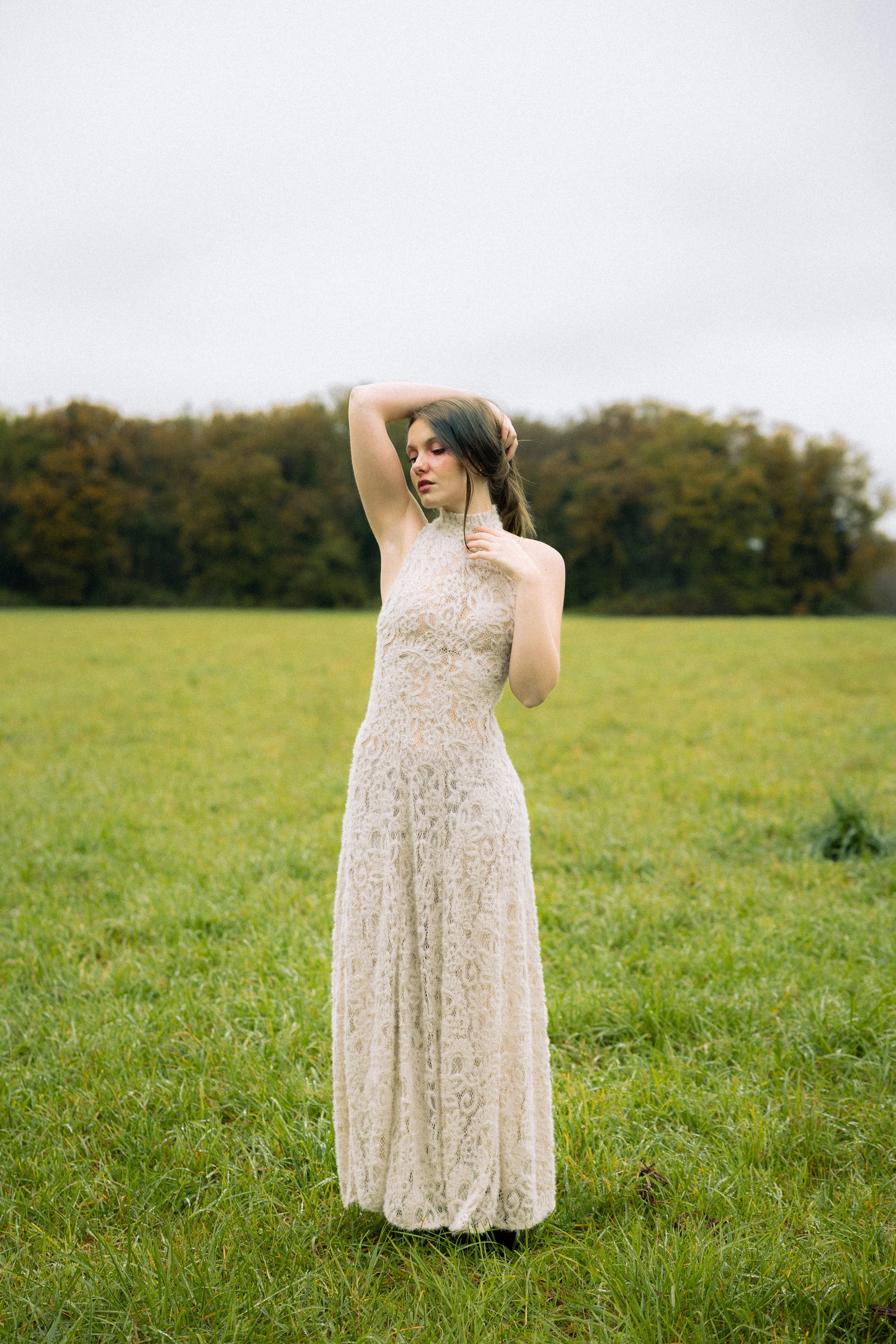 Une jeune femme portant une robe longue en dentelle beige, debout dans un champ verdoyant avec des arbres en arrière-plan, sous un ciel gris.
