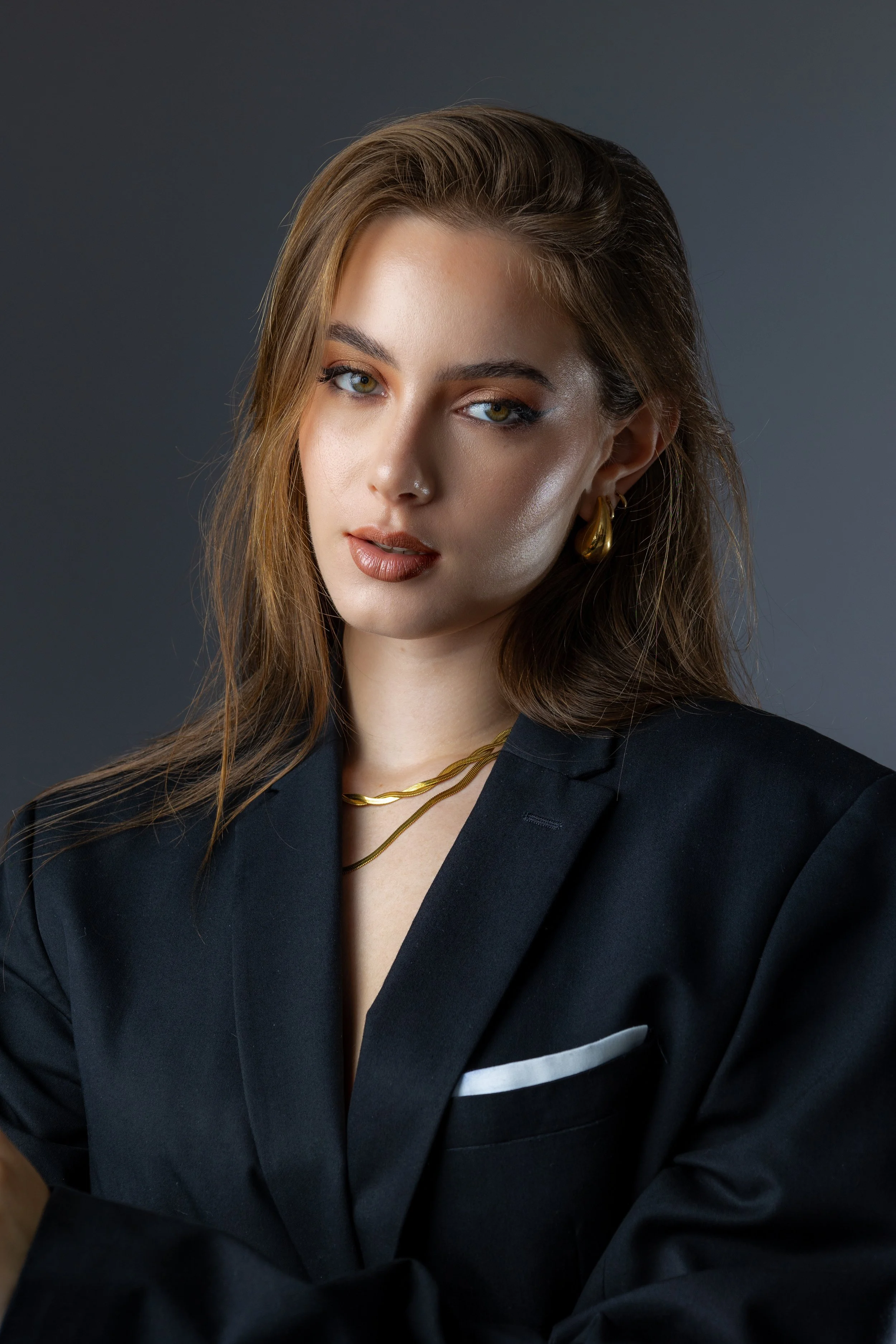 Jeune femme avec des cheveux bruns, portant un blazer noir et des bijoux en or, regardant vers la caméra avec un léger sourire contre un fond gris