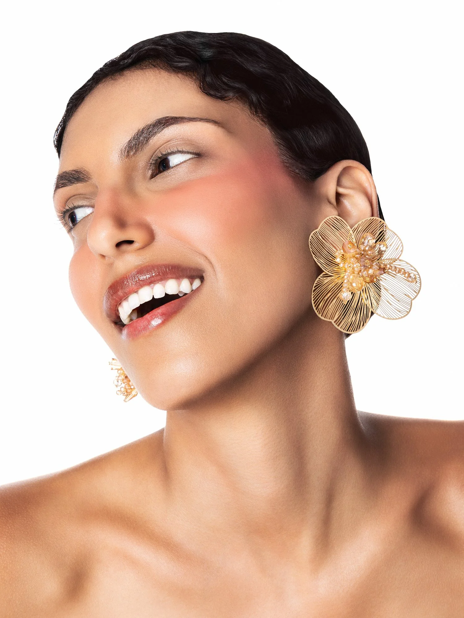 Portrait d'une femme avec de grandes boucles d'oreilles dorées en forme de fleur, maquillage neutre, souriant légèrement, fond blanc.