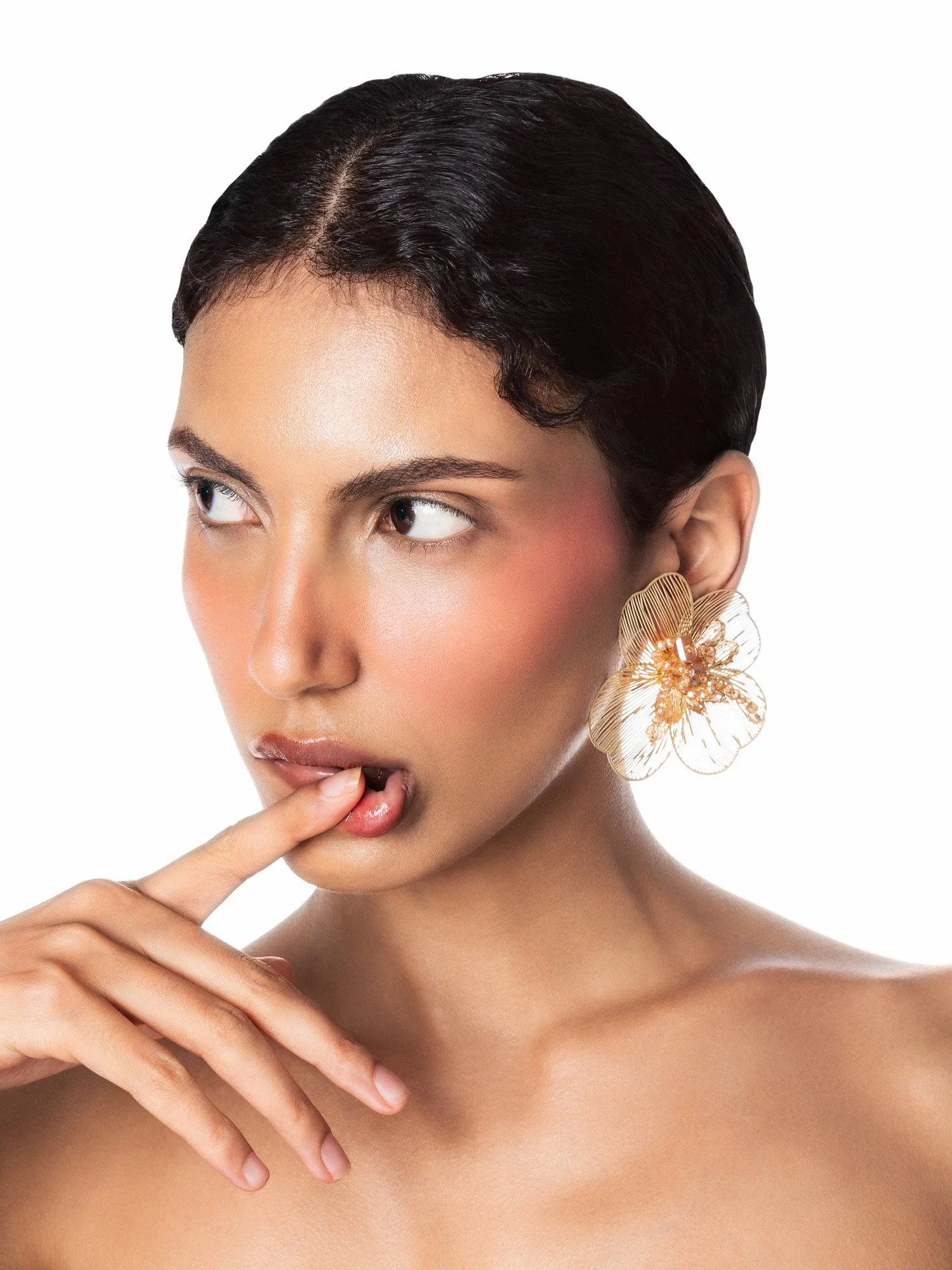 Une femme avec une grosse boucle d'oreille en forme de fleur flottante, touches maquillage naturelles, yeux qui regardent de côté, et doigt près de la bouche, fond blanc.