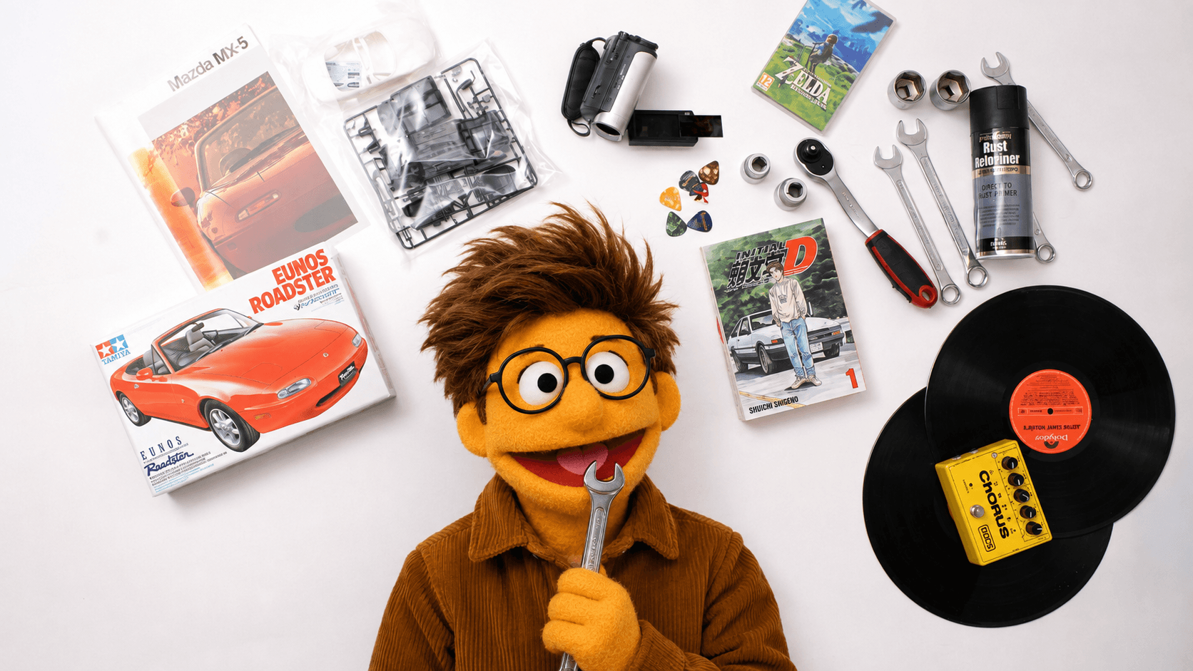 A person dressed as a puppet from Sesame Street with brown hair, yellow face, and a wide smile standing in front of a red convertible sports car on a road surrounded by green trees.