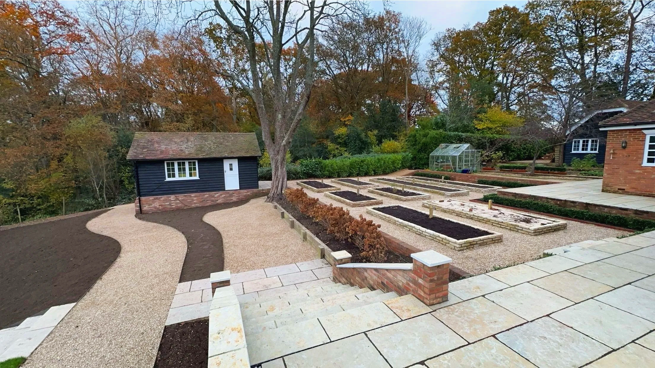 A backyard garden with a paved patio, layered garden beds, a gravel pathway, a shed, a greenhouse, and mature trees with autumn foliage.