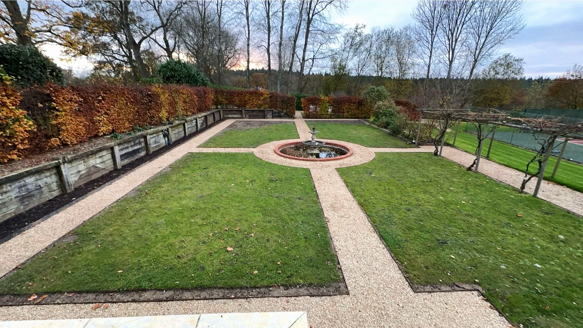 A landscaped garden with a central fountain surrounded by gravel paths, green grass patches, and a hedge on the left side, with trees in the background during late fall.