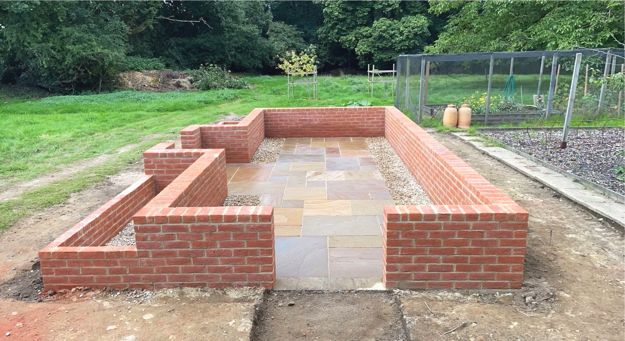 A small backyard patio with brick walls and a stone tile floor, surrounded by grass and garden beds.