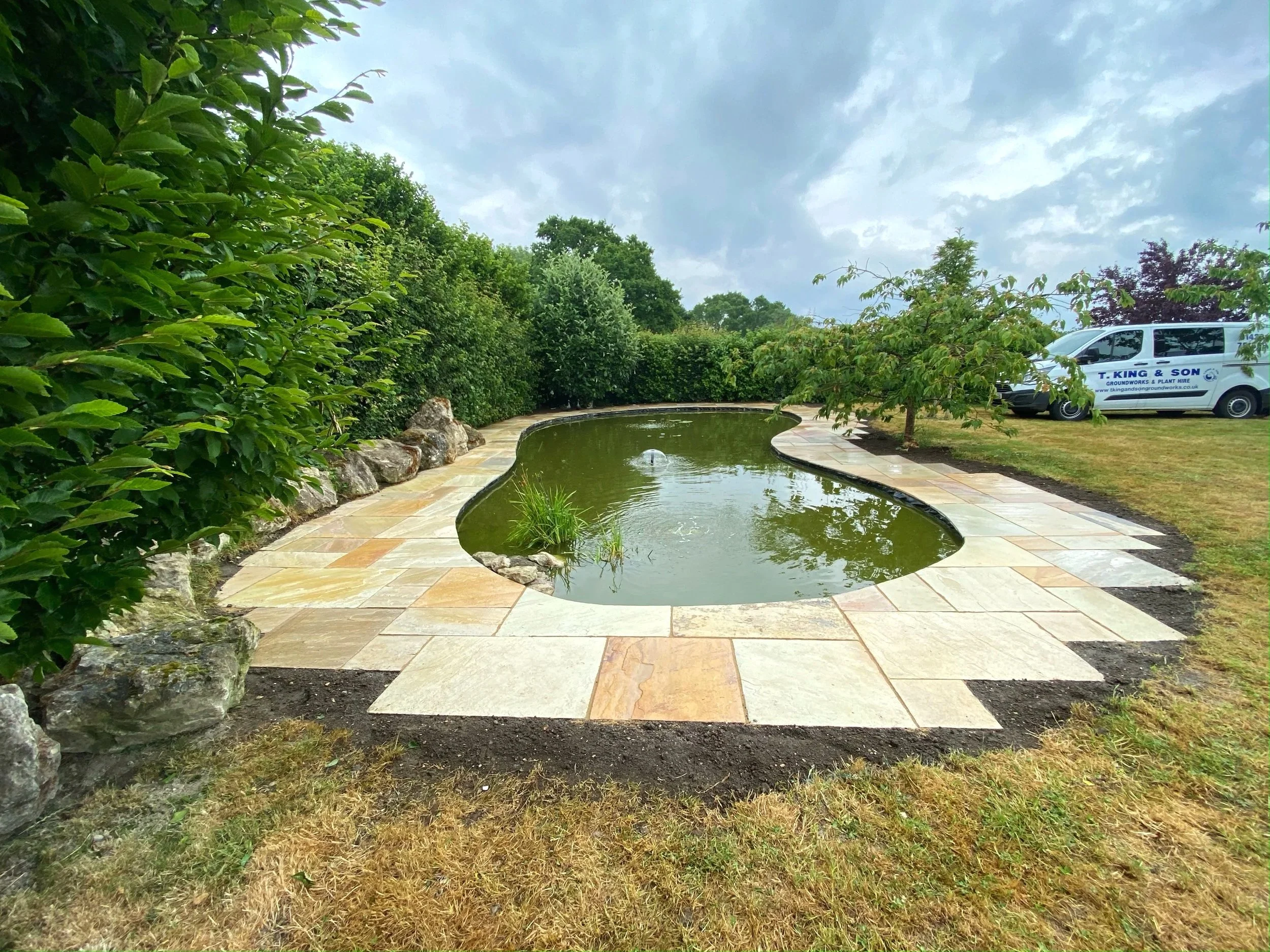Small kidney-shaped pond with greenery on the edges, surrounded by a stone-paved walkway, in a garden area with trees and bushes, and a white van parked nearby under an overcast sky.