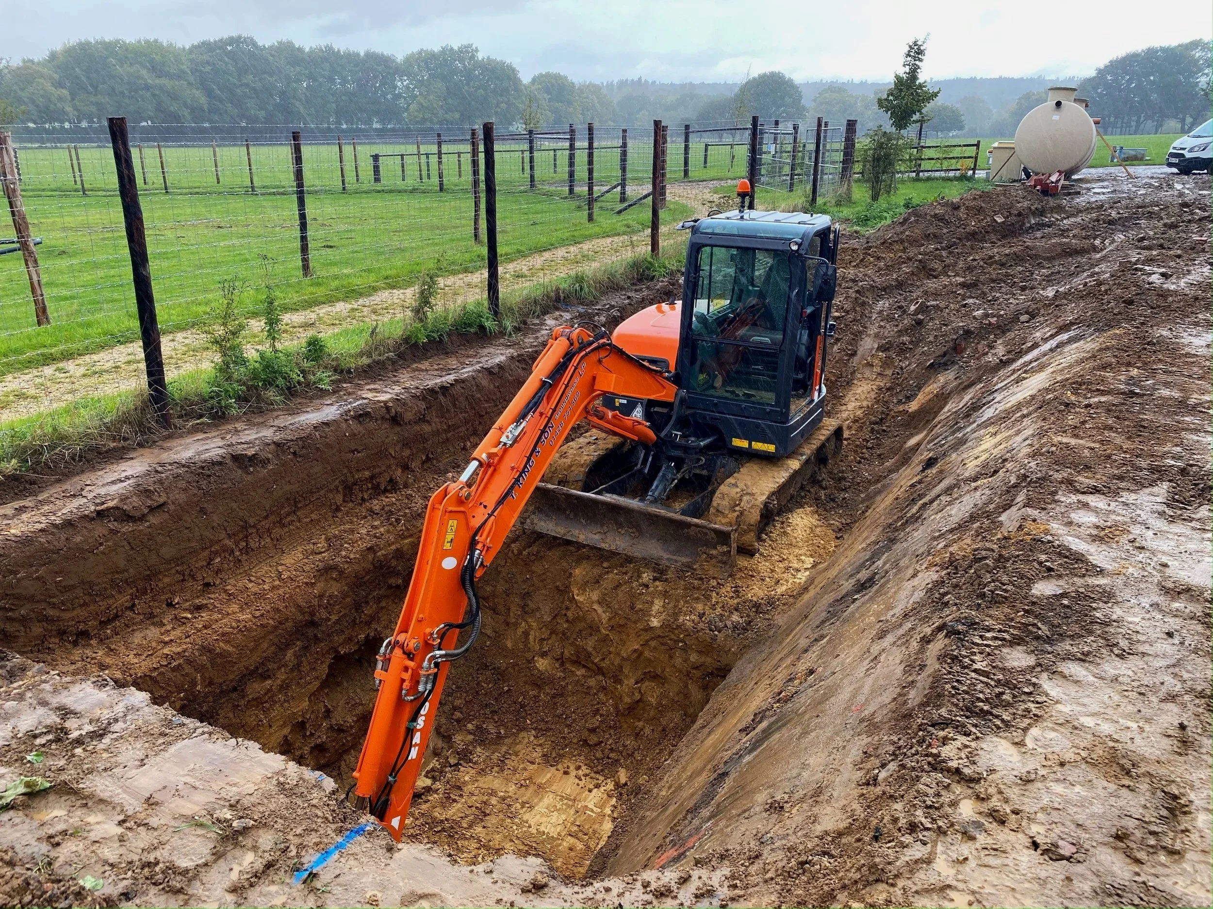 An orange excavator working in a large trench on a construction site with a fence, green fields, and trees in the background, and a white tank and vehicle nearby.