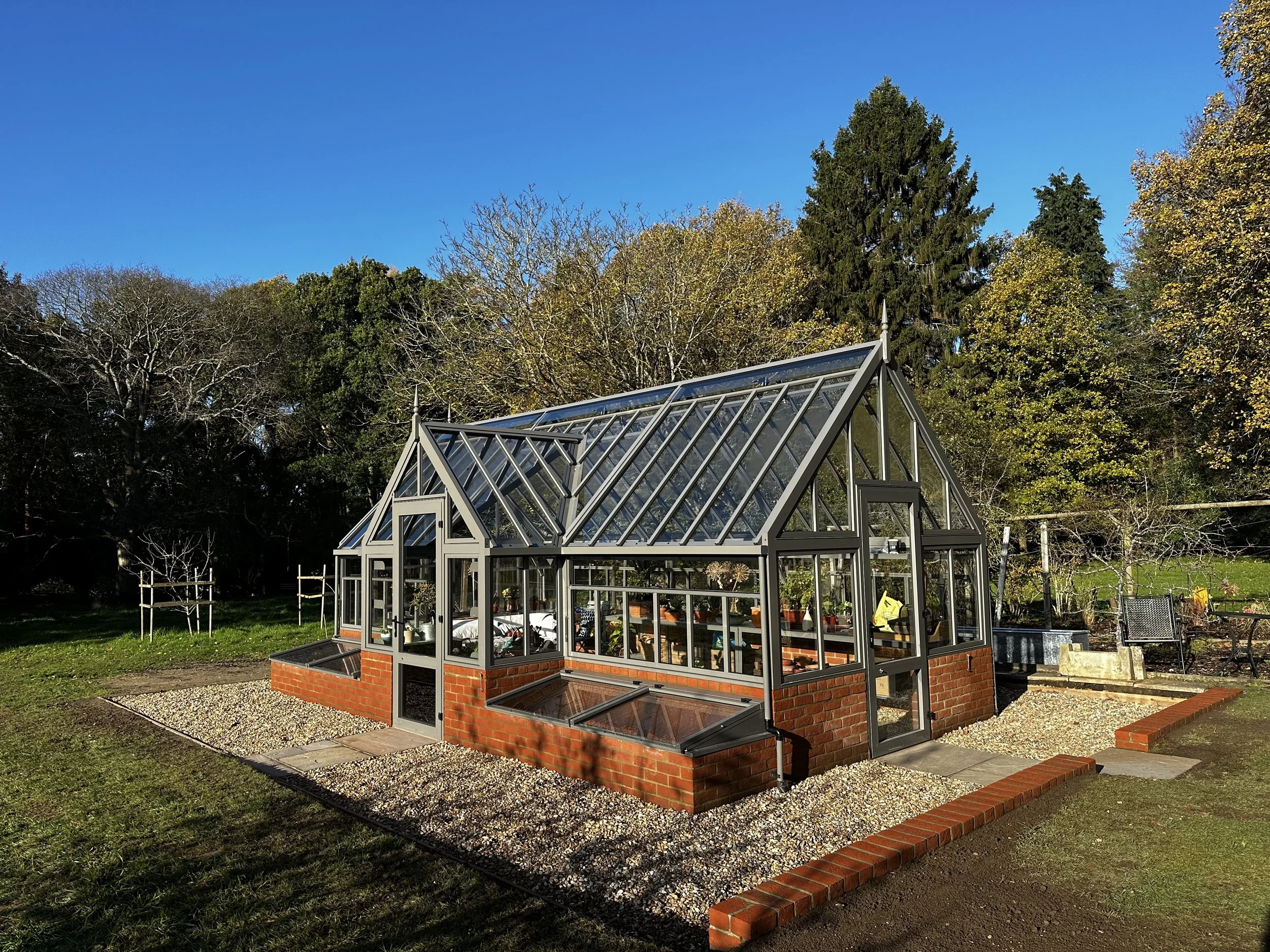 A greenhouse with a brick base and metal framing, situated in a garden with trees and a blue sky in the background.