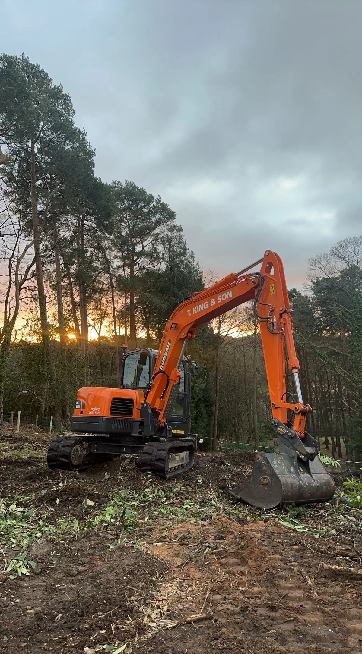 An orange excavator with the words 'T. King & Son' on its arm, working on a dirt site with trees in the background at sunset, under a cloudy sky.