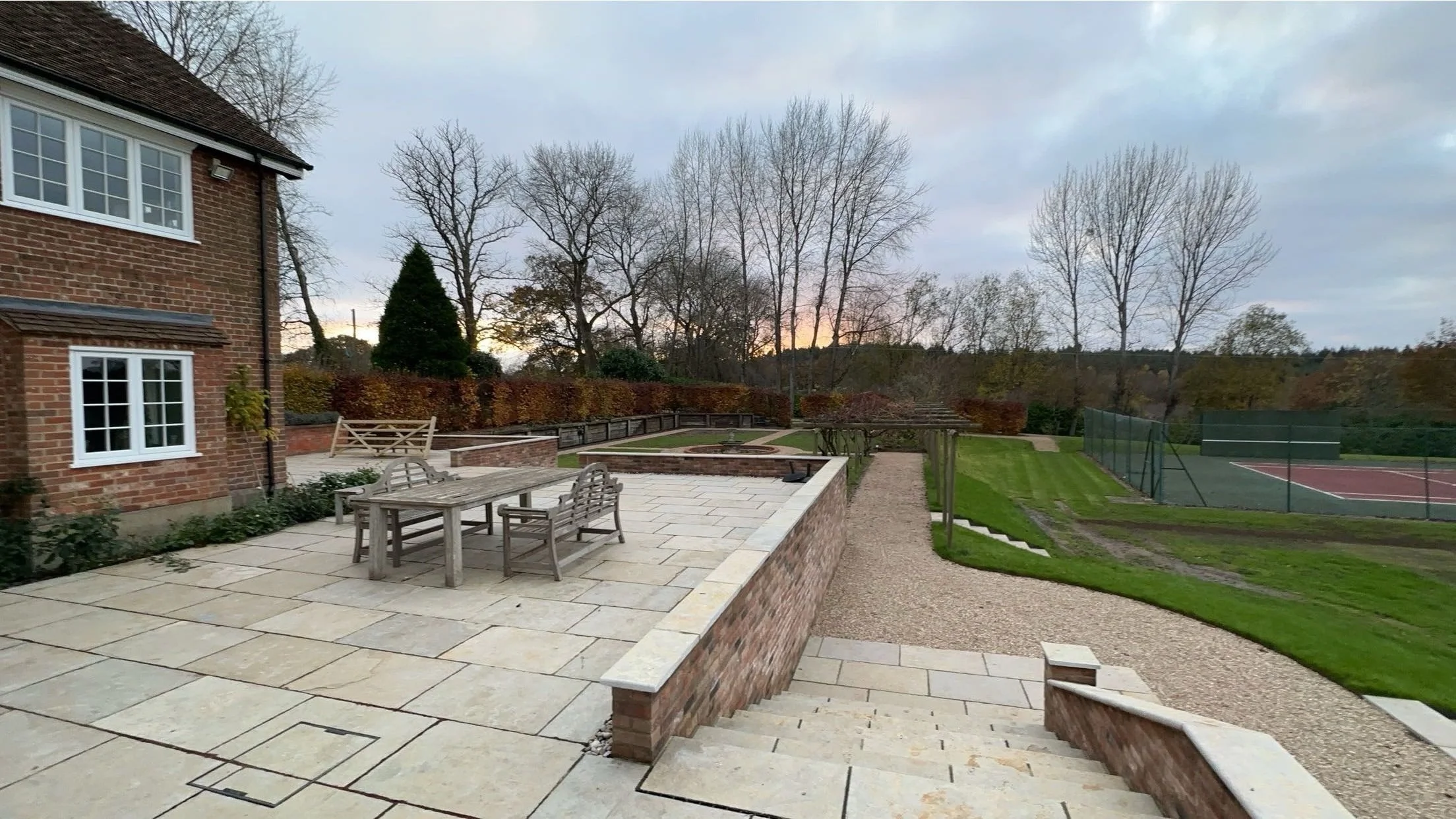View of a patio with outdoor furniture, stairs, a gravel pathway, and a tennis court in a backyard with trees and a cloudy sky at sunset.