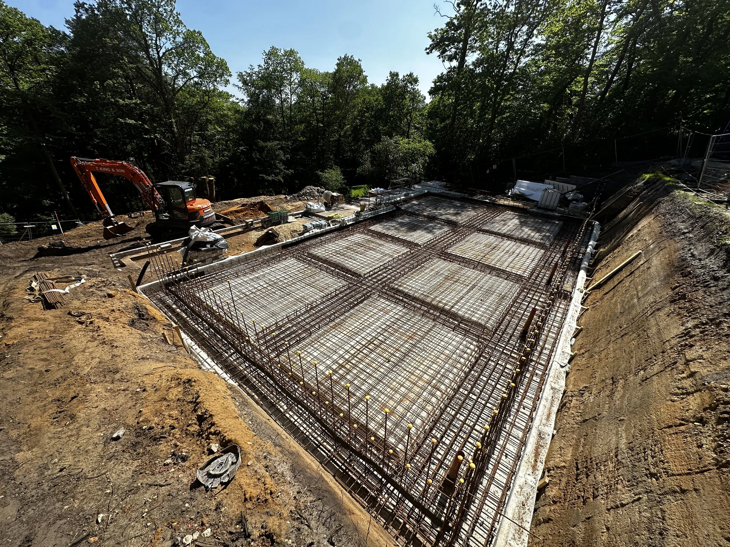 Construction site with steel rebar framework for a building foundation, surrounded by dirt and construction equipment, in a wooded area.