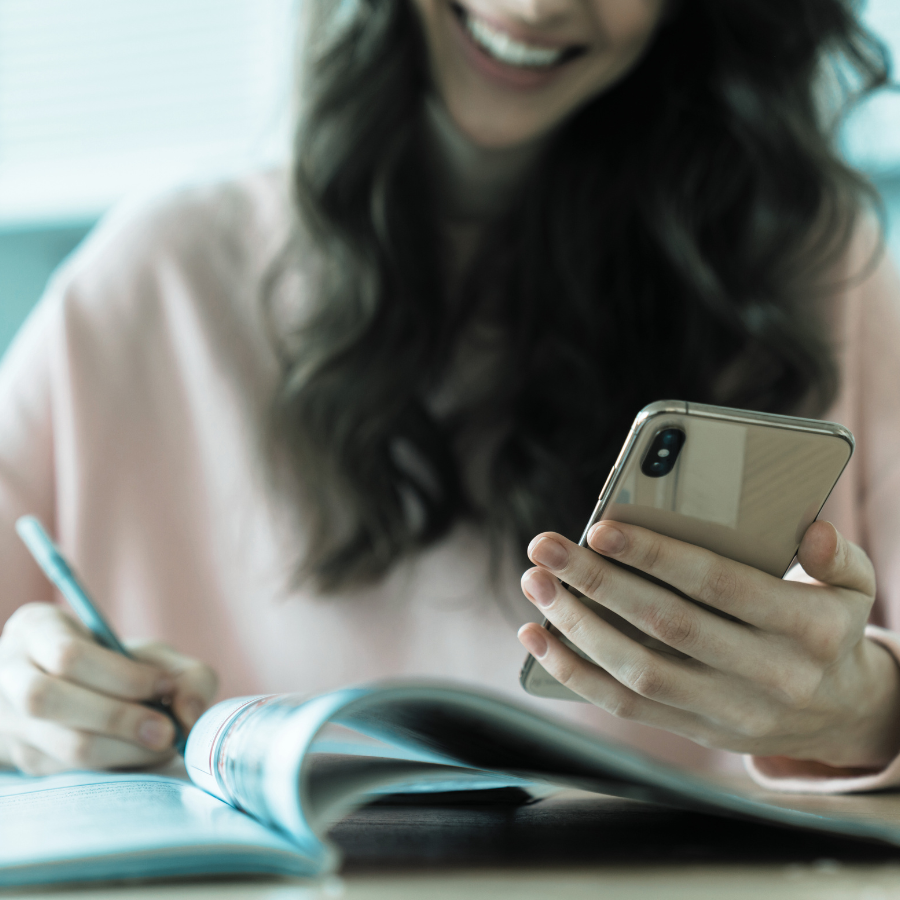 A woman with long dark hair smiling while looking at her smartphone, with an open notebook in front of her, in a bright indoor setting.