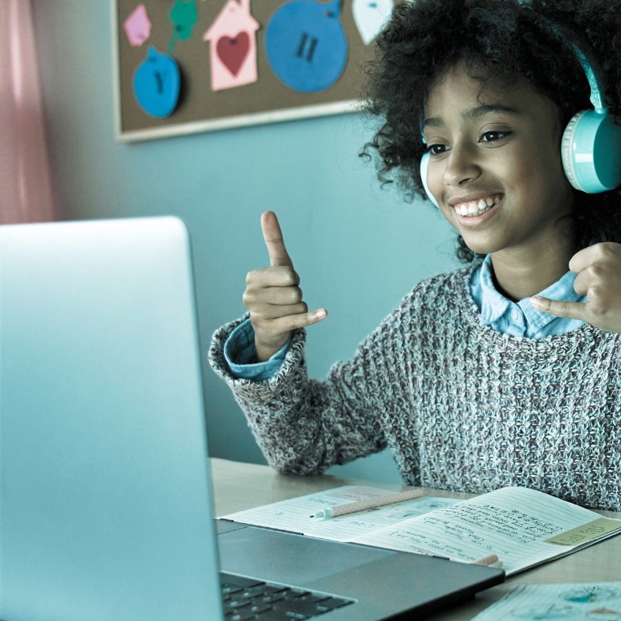 A young girl with curly hair wearing a sweater and headphones smiling and giving a thumbs-up while looking at her laptop in a classroom or study area.