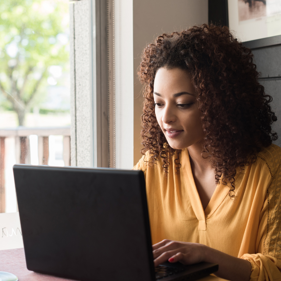 Young woman with curly hair working on a laptop at a table near a window with green trees outside.