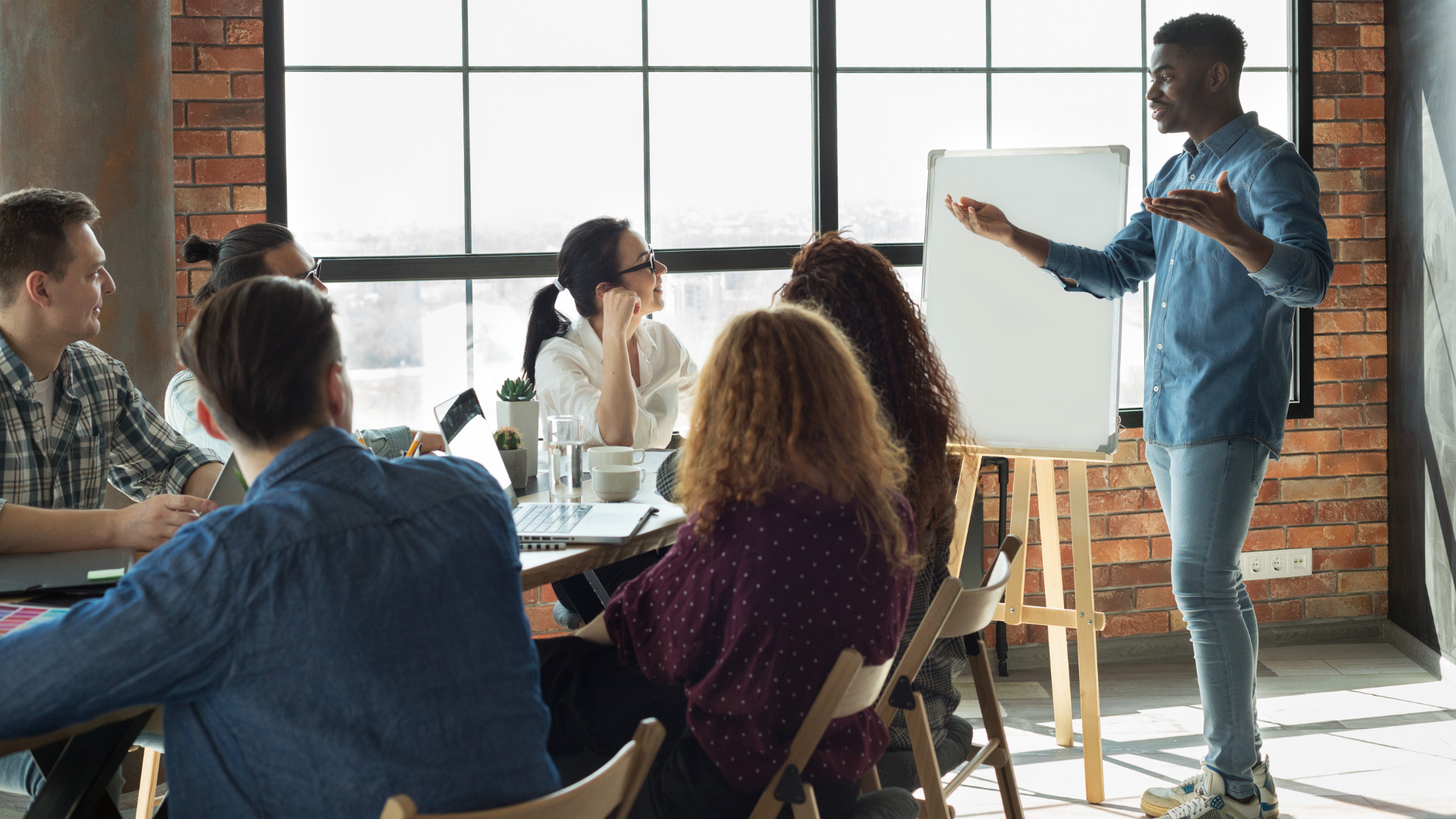 A black man leads a meeting, standing and presenting to a homogenous looking group of people