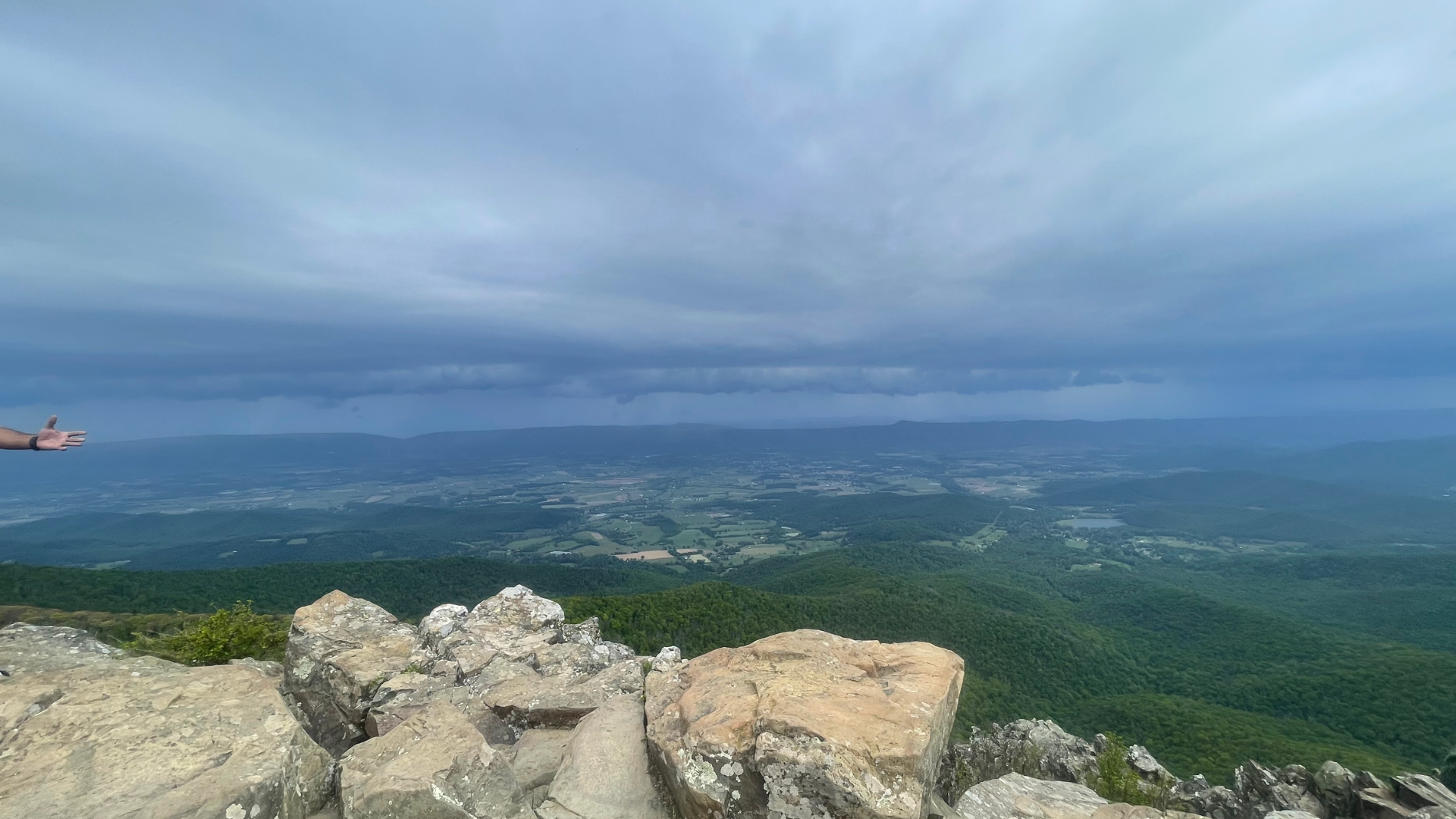 A wide panoramic scenic view looking out from a rocky mountain peak under a dramatic, cloudy sky.