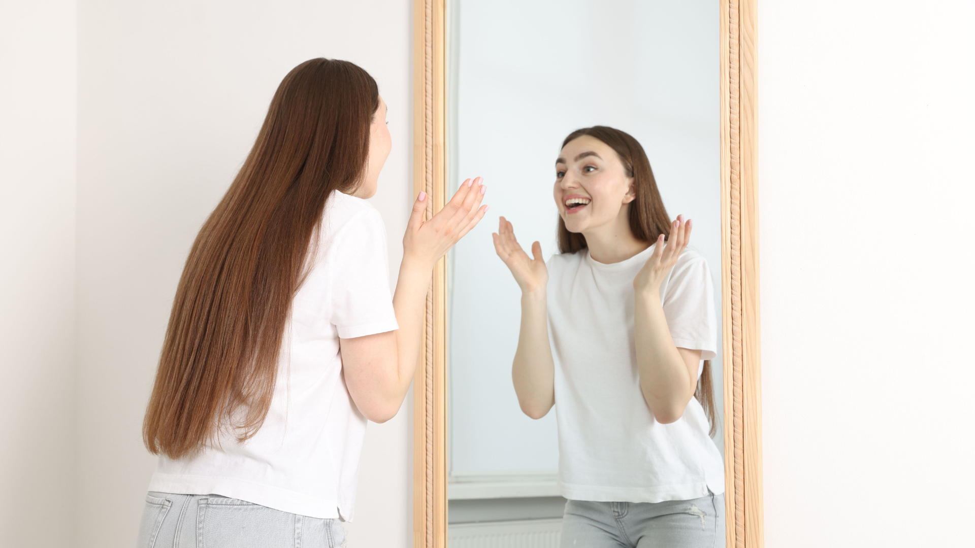 Young woman with long brown hair smiling and talking to her reflection in the mirror.