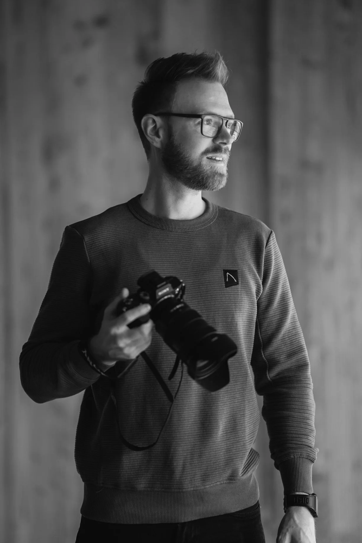 Ein Mann mit Brille, Bart und Kurzhaarfrisur hält eine Kamera in der Hand, blickt nach rechts und trägt eine Armbanduhr sowie einen Pullover.
