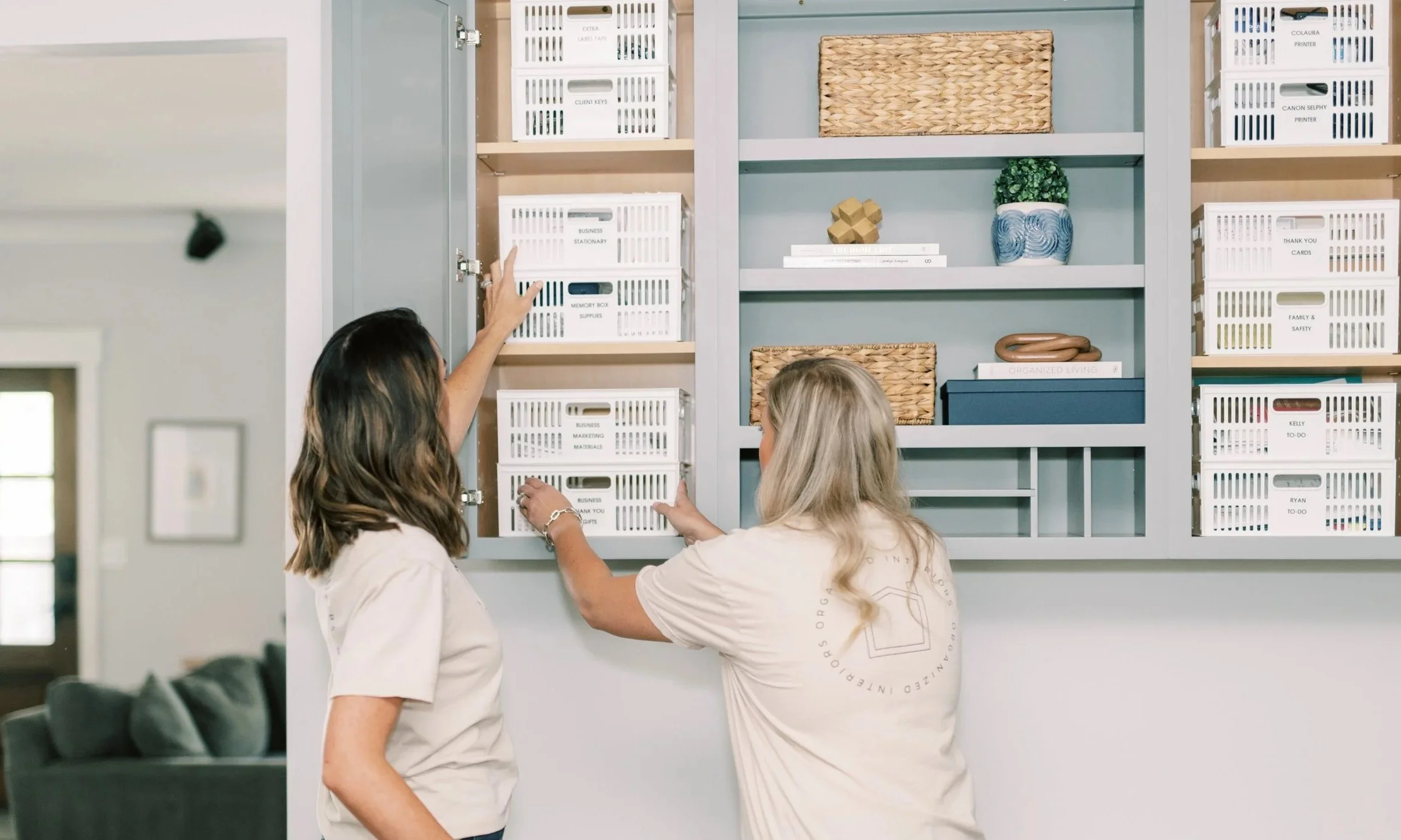 Two professional organizers arranging labeled storage bins inside a custom home office cabinet to create an organized and functional workspace.