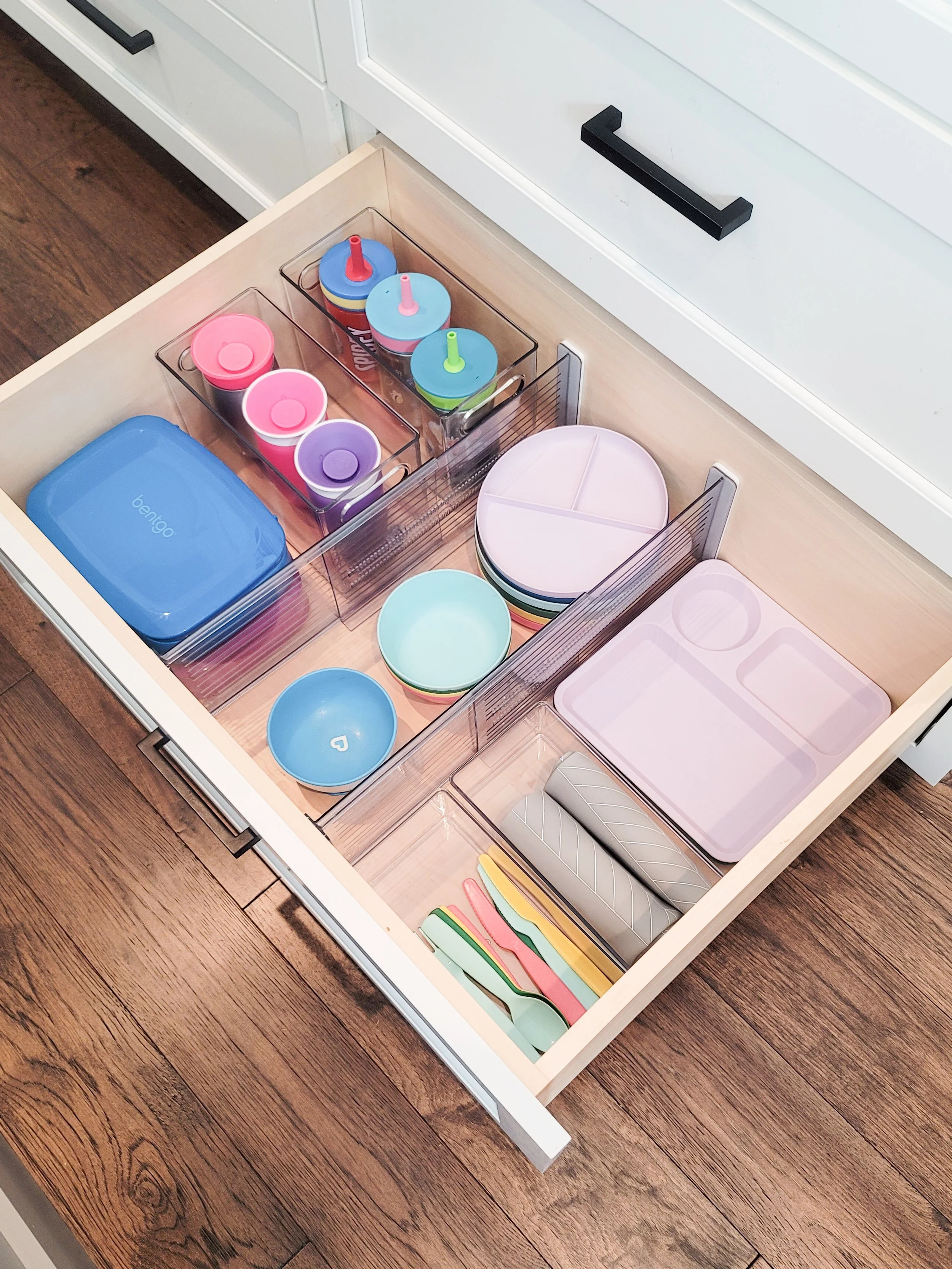 Organized kitchen drawer with clear storage bins holding kids' lunch containers, reusable cups, plates, bowls, and utensils for easy meal prep.
