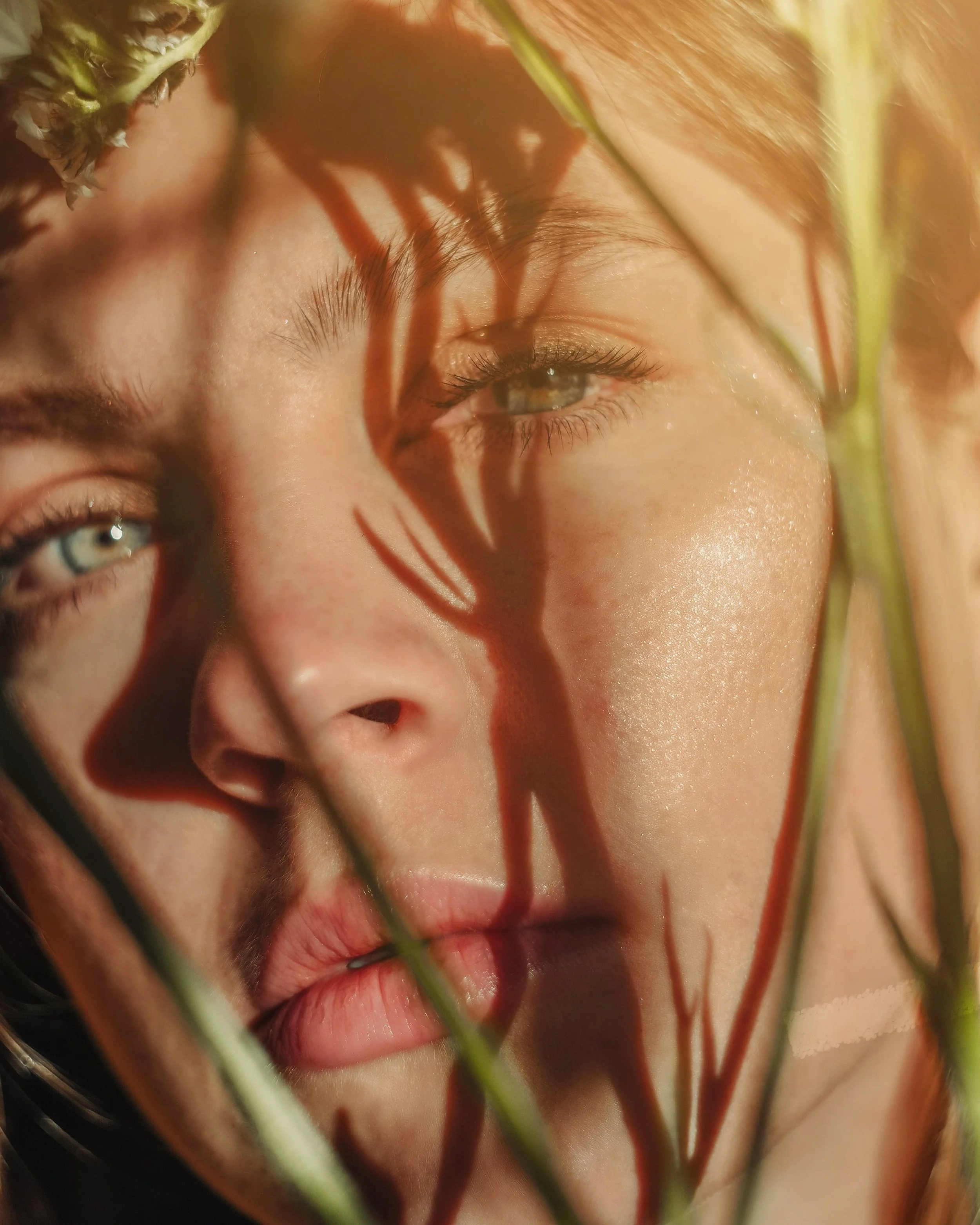 Close-up of a woman's face with shadows of plant stems and leaves cast on her skin, highlighting her green eyes and facial features.