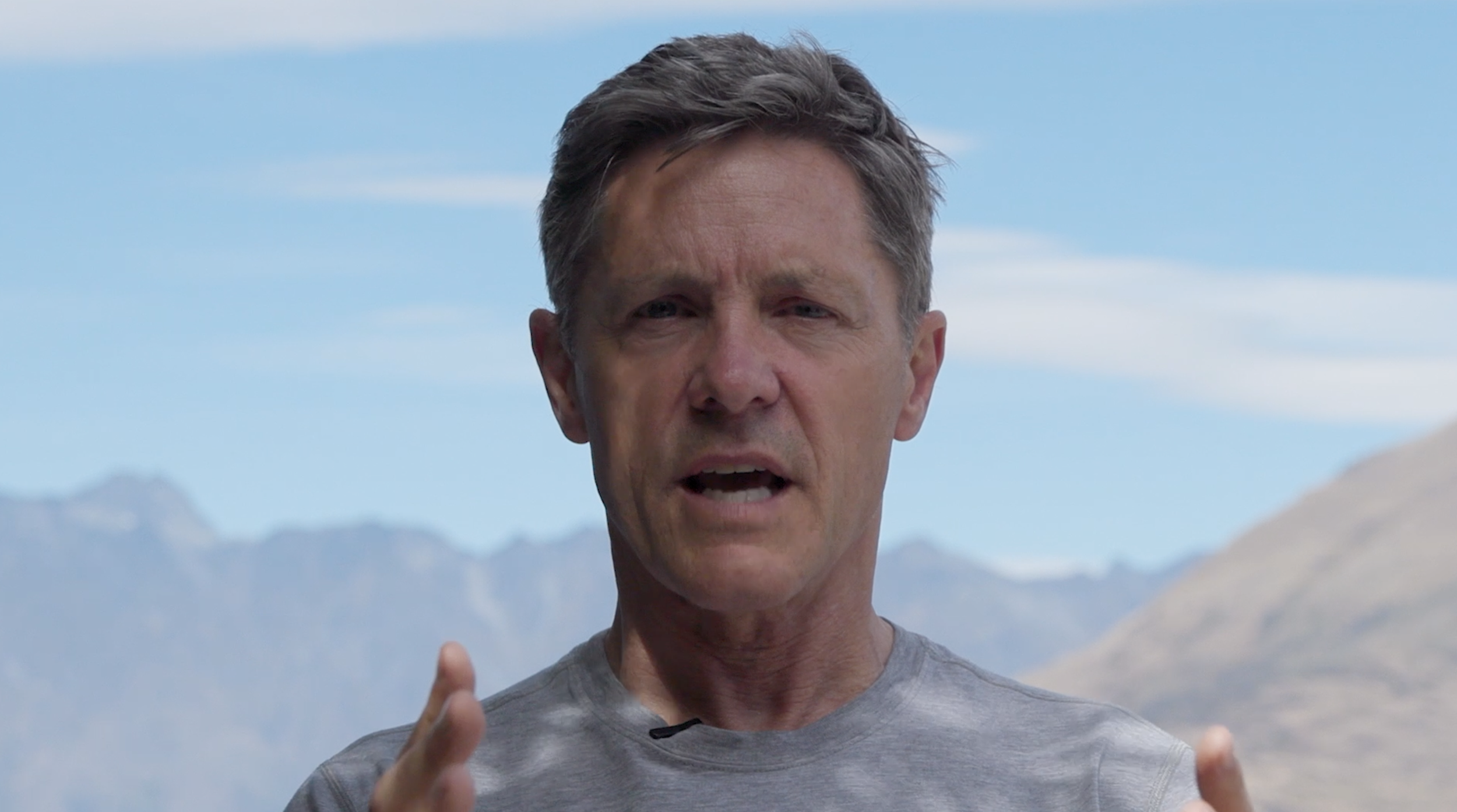 A middle-aged man with short, gray hair speaking outdoors in front of a mountain range and blue sky, wearing a gray athletic shirt.