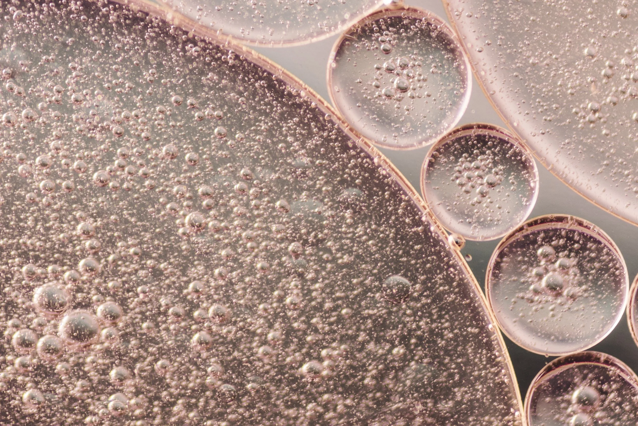 Close-up of oil and water droplets in a thin film, with tiny bubbles and circular patterns in a pinkish hue.