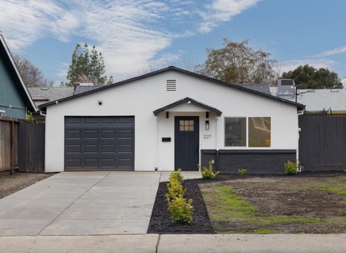 A modern single-story house with a white exterior, dark gray garage door, and front door, with a concrete driveway and a small landscaped yard with young bushes.