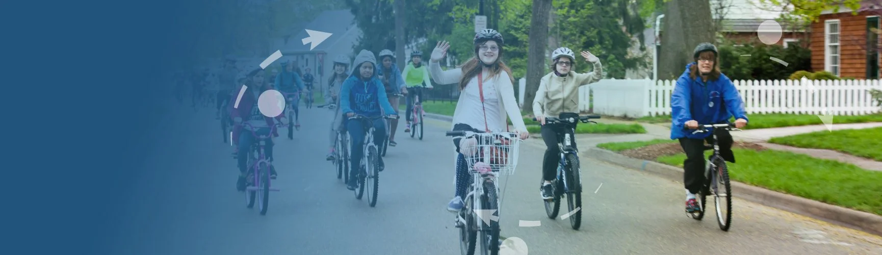 Group of children and adults riding bikes on a suburban street lined with trees and houses, with one girl smiling and waving at the camera.