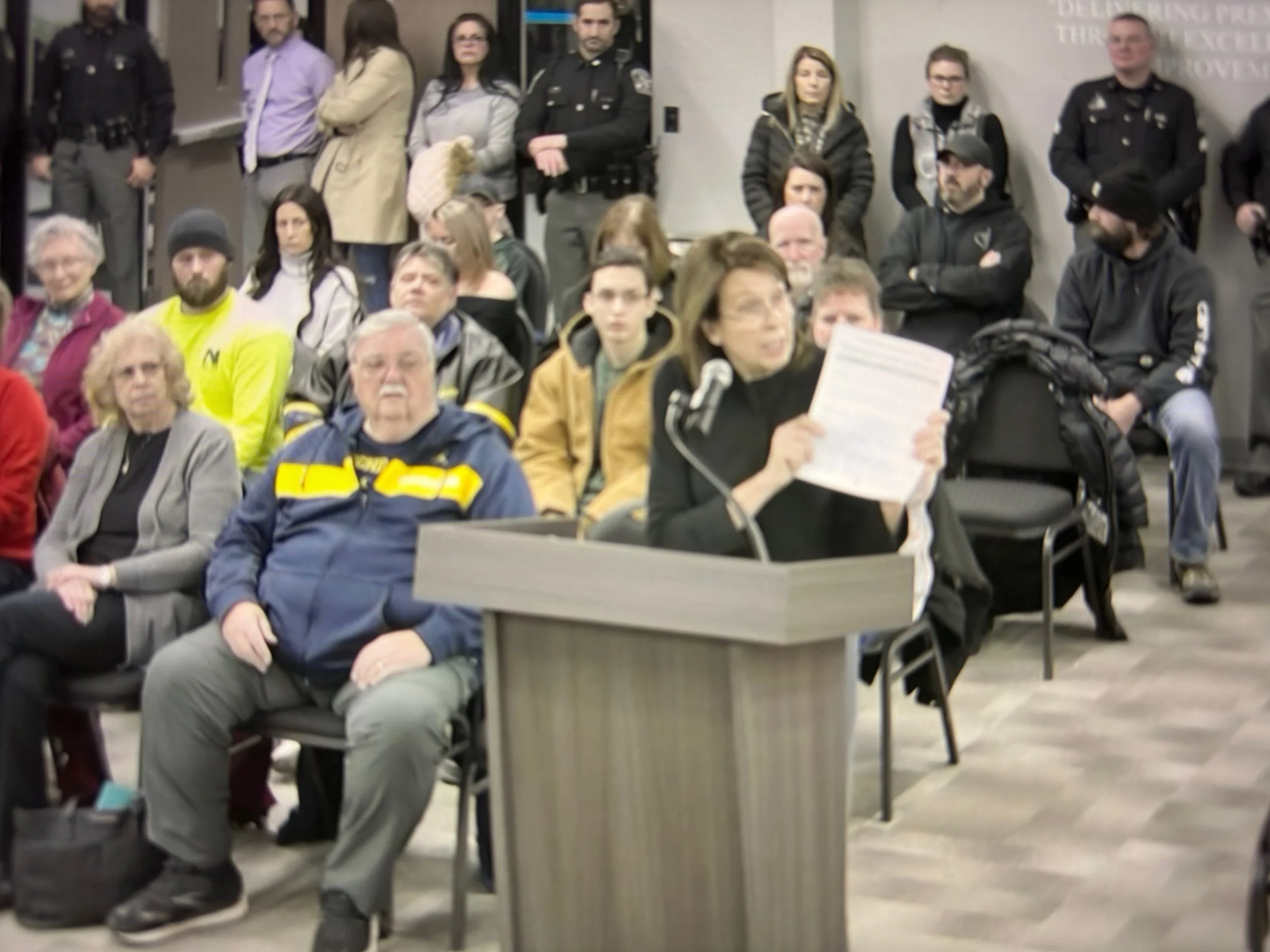 Woman speaking at a podium during a public meeting or hearing, holding a paper and reading aloud while an audience listens.