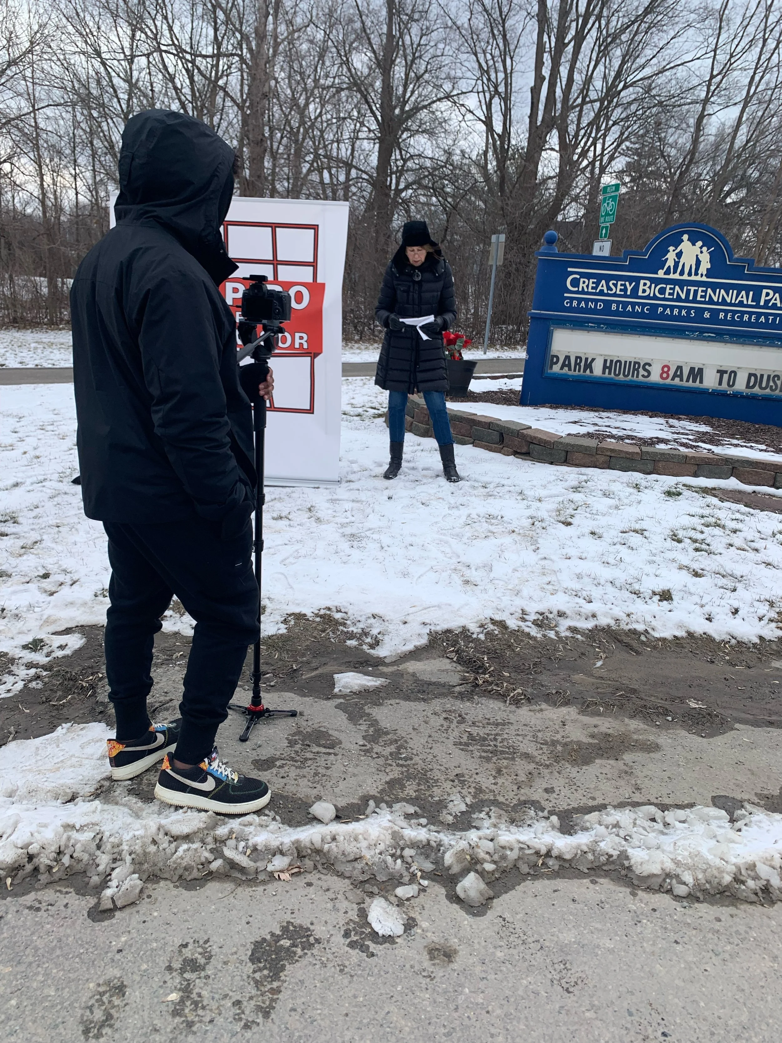 A person in a black jacket with a hood filming another person speaking outdoors at Creasey Bicentennial Park in winter, with snow on the ground and a blue sign indicating park hours from 8 am to dusk.