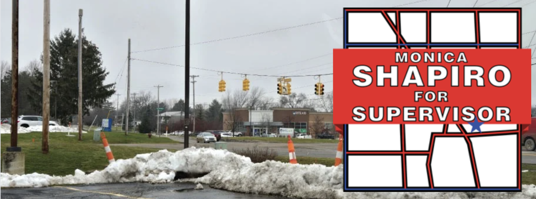 A road intersection with traffic lights, cars, and a building in the background, with a large campaign sign in the foreground supporting Monica Shapiro for Supervisor.