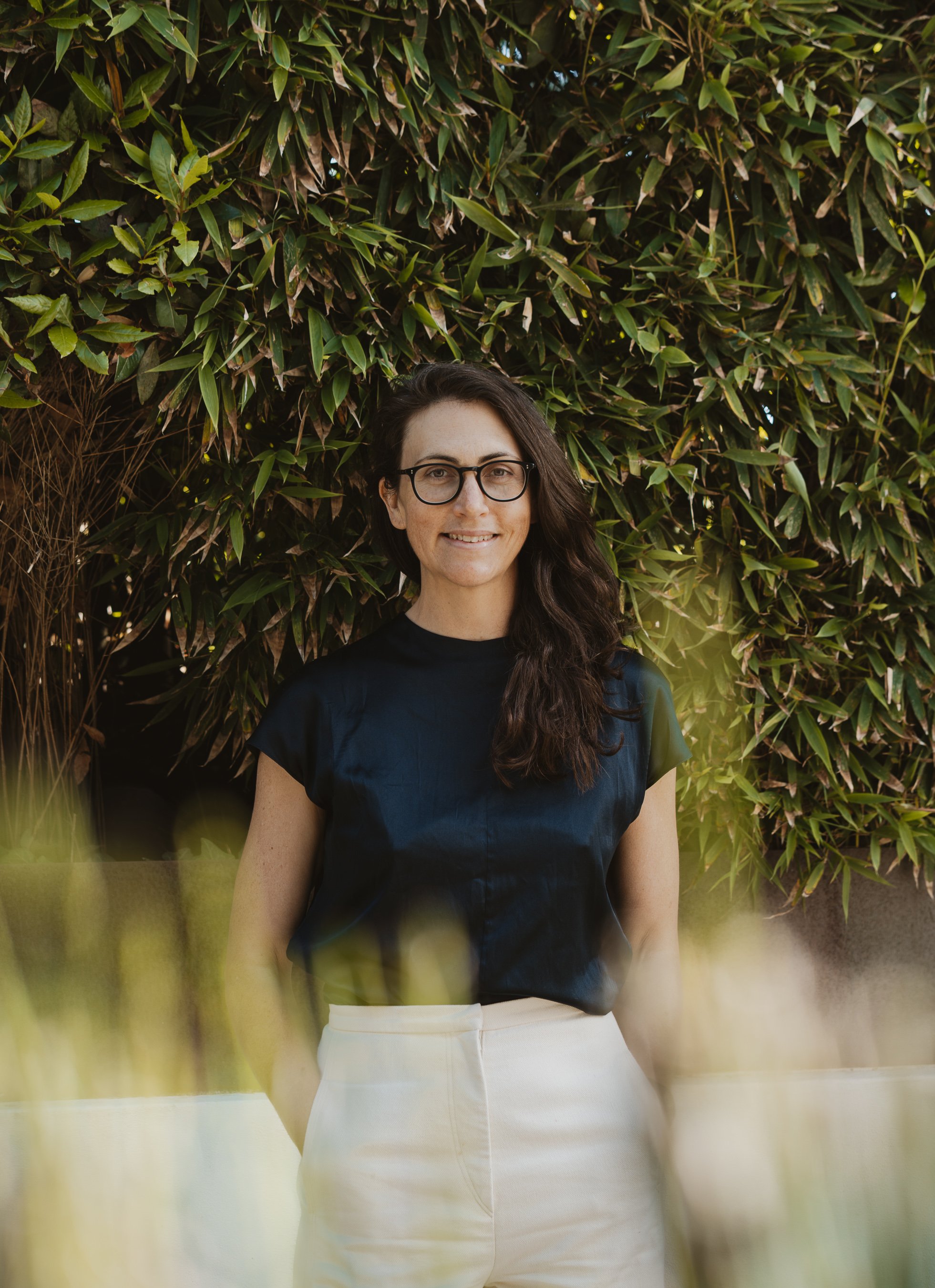 A woman with long dark hair, wearing glasses, a dark blue top, and a white skirt, standing outdoors in front of green leafy bushes, smiling at the camera. Maggie Spicer Biarritz