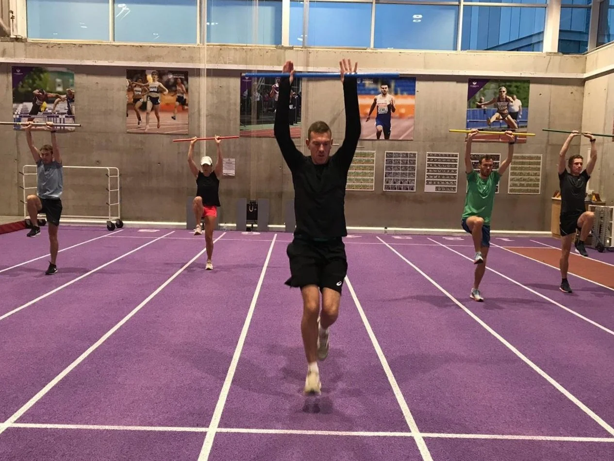 Multiple athletes practicing sprint drills on a purple indoor track, with posters of runners on the wall behind them.