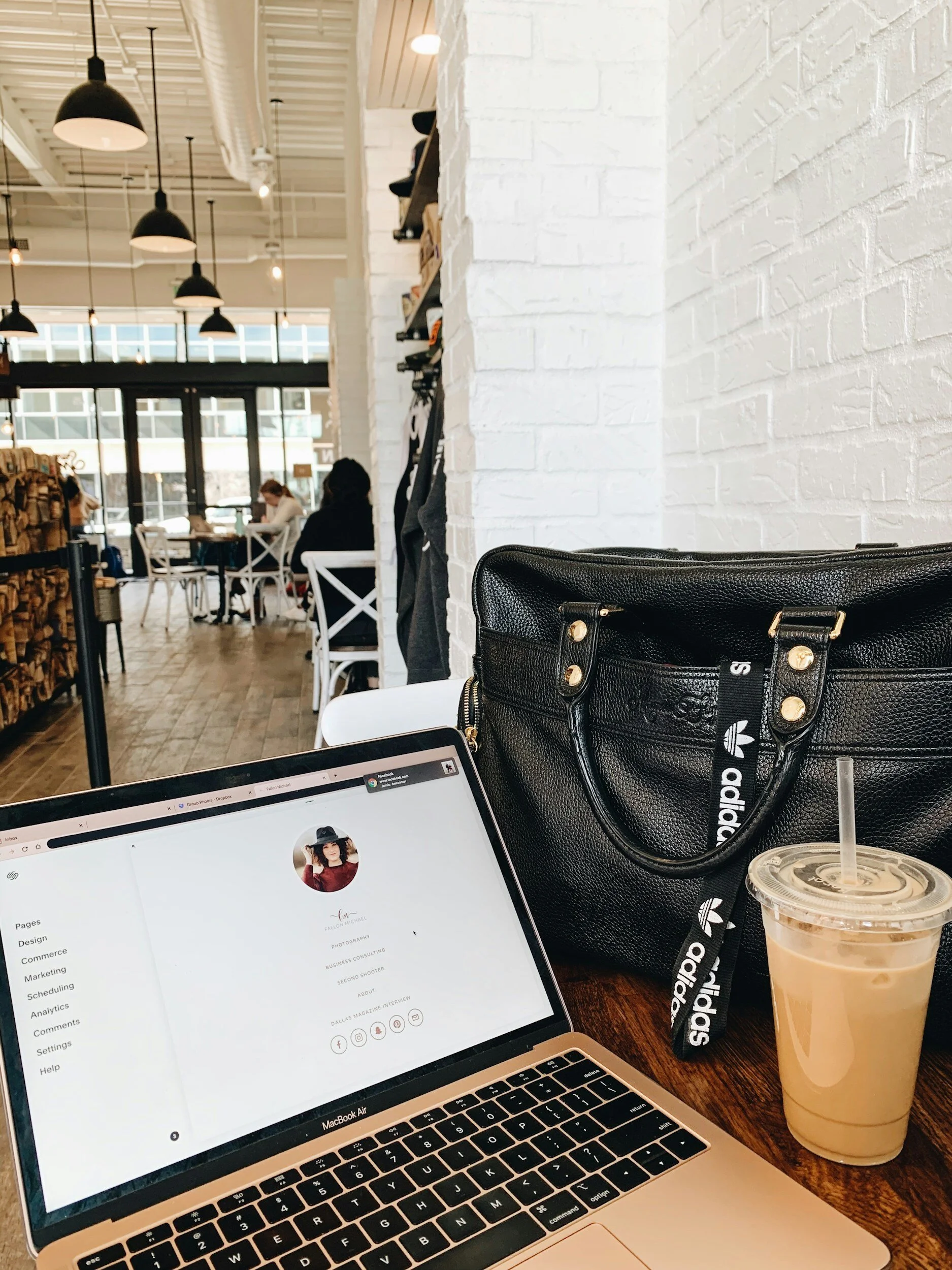 A laptop on a table in a coffee shop, showing a website profile page, with a black leather purse, iced coffee, and a lanyard reading 'adidas' nearby. The cafe has white brick walls, wooden floors, and pendant lighting, with customers seated in the background.