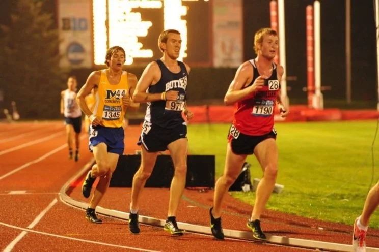 Three male runners competing in a track race at night, on a red clay track with a digital scoreboard and stadium lights in the background.