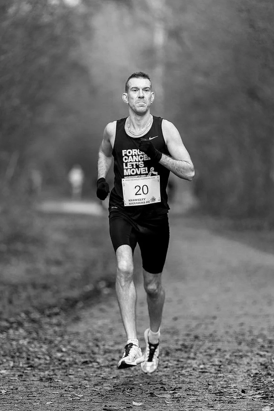 A man running on a trail wearing a tank top with a message about cancer awareness, black shorts, gloves, and a race bib with number 20.